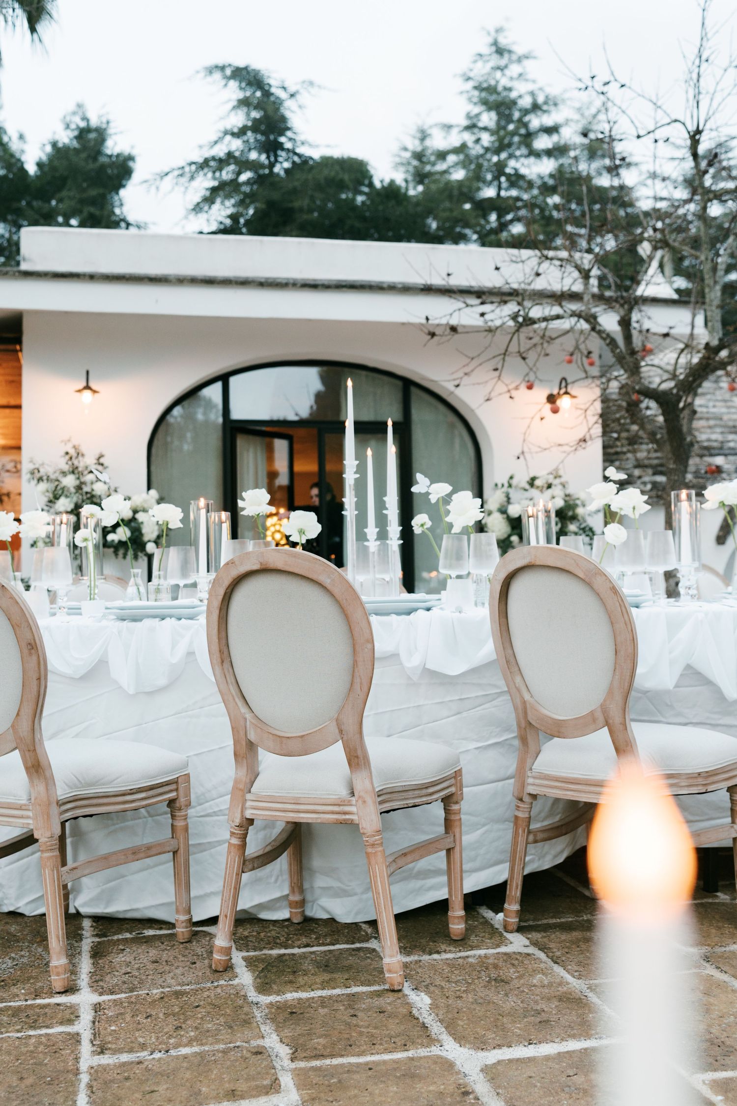 Outdoor wedding reception table with white flowers, tall candles, and Louis XVI chairs at dusk. Puglia wedding photographer,