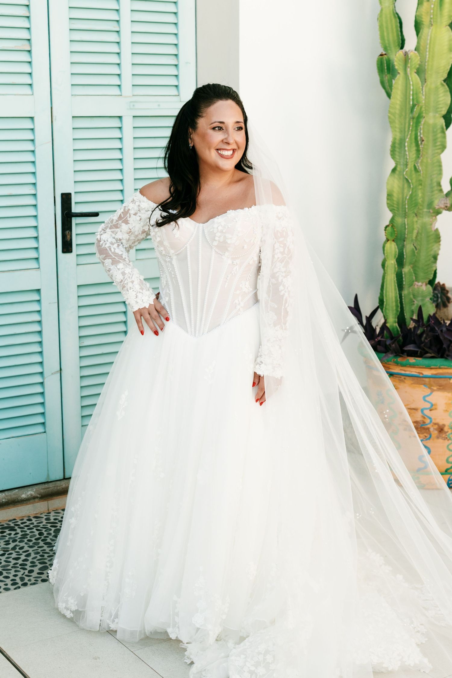 Smiling bride in an off-the-shoulder lace A-line wedding gown with a corset bodice and long veil, posing by a teal door and cactus.