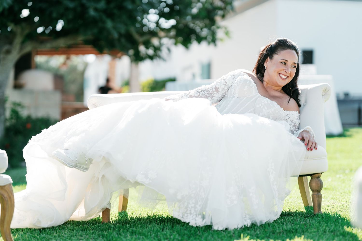 Smiling bride in a white lace wedding gown with a full tulle skirt reclines on a wooden chair on a sunny green lawn.