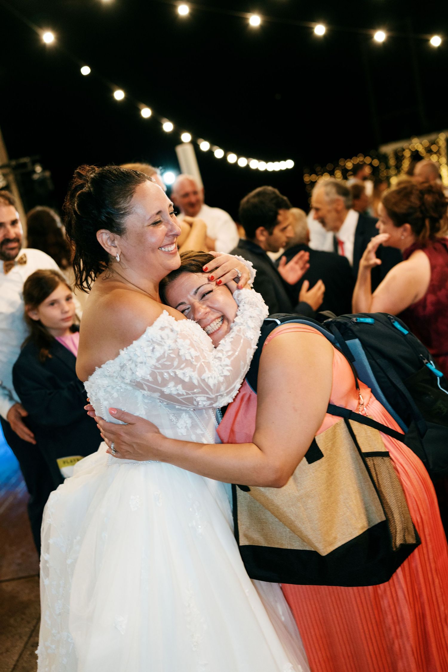 Bride in a white lace wedding gown shares a joyful embrace with a smiling guest at an evening reception under string lights.