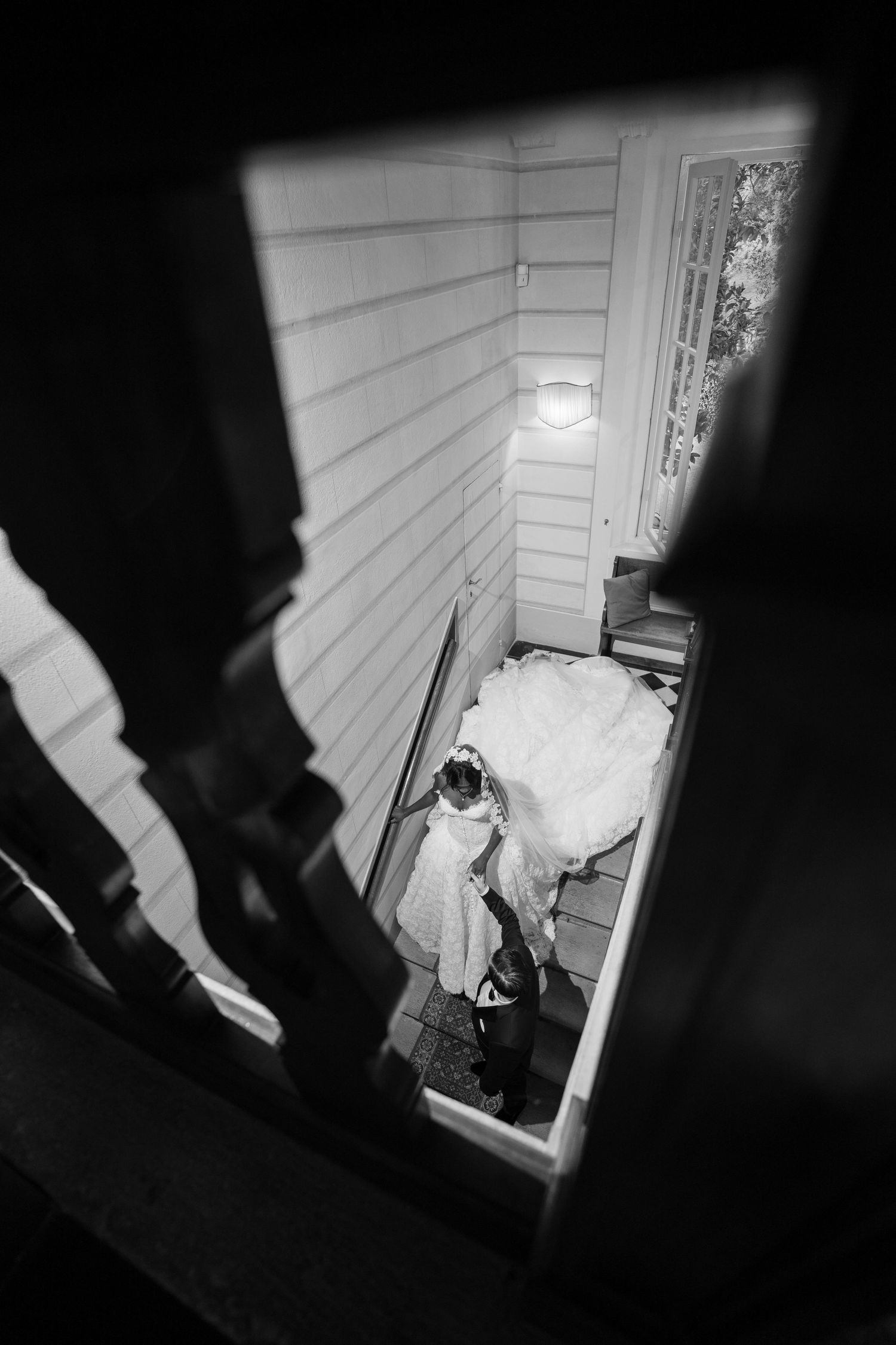Black and white overhead photo of a bride in a flowing wedding gown with a long train standing on a narrow staircase beside a groom.