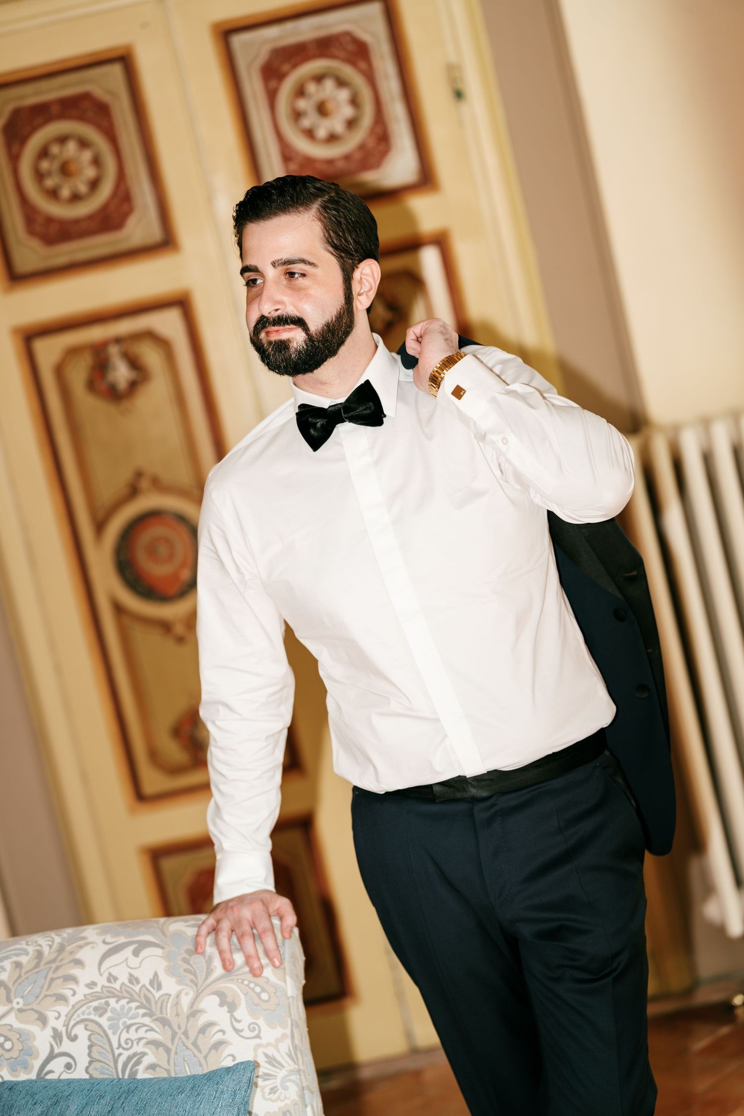 Bearded groom in a white dress shirt and black bow tie holds his tuxedo jacket over his shoulder in an elegant room with ornate panels.