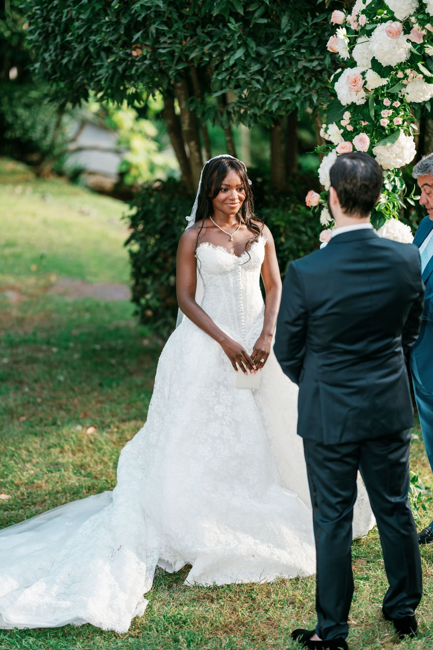 Bride in a strapless white lace wedding gown and veil faces her groom during an outdoor garden ceremony beside pink and white roses.