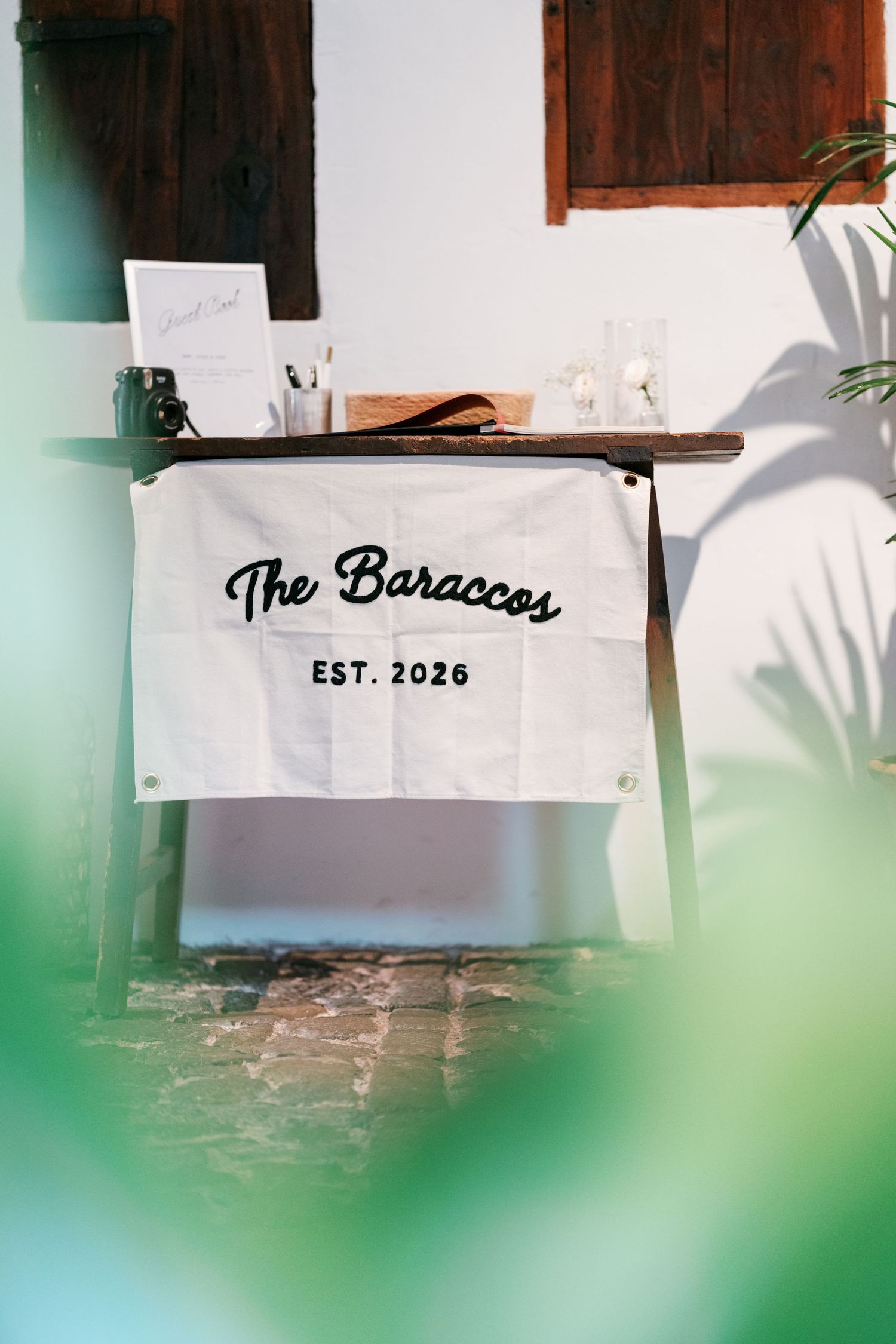 Cream fabric banner reading The Baraccos Est. 2026 hangs from a rustic wooden table with wedding décor, framed by blurred green leaves.