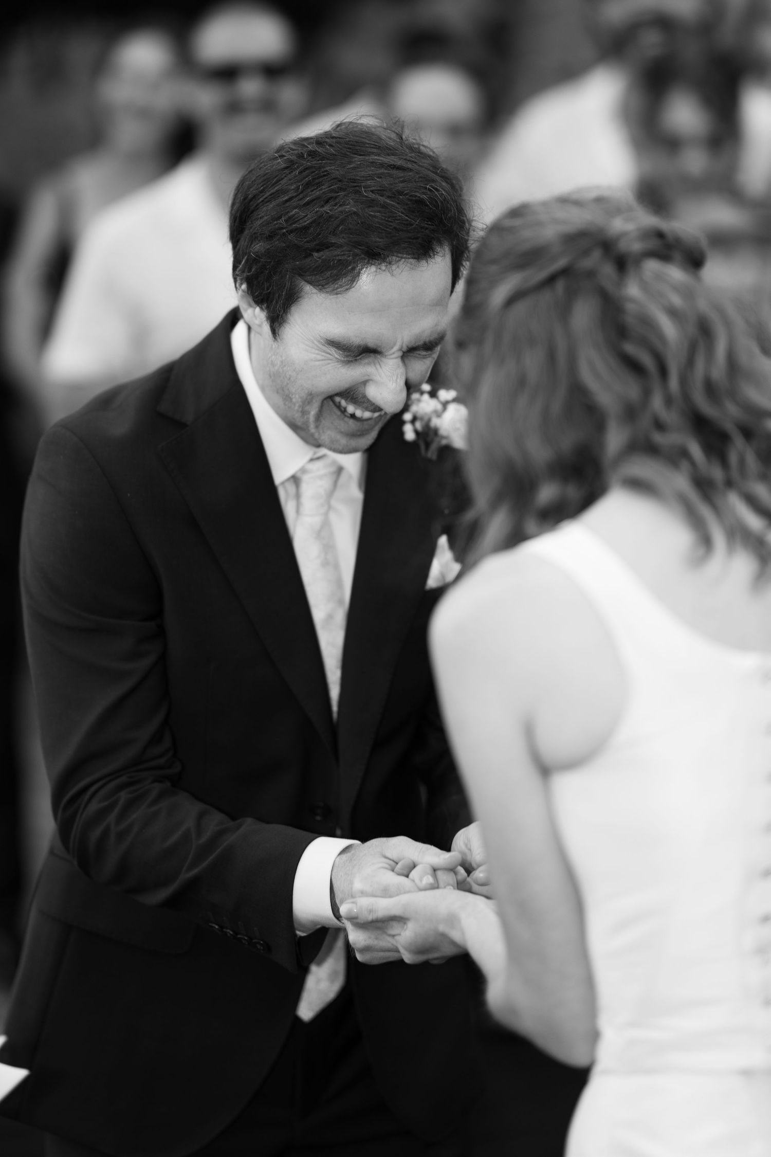 Smiling groom in a dark suit exchanges rings with his bride during an outdoor wedding ceremony, black and white photo.