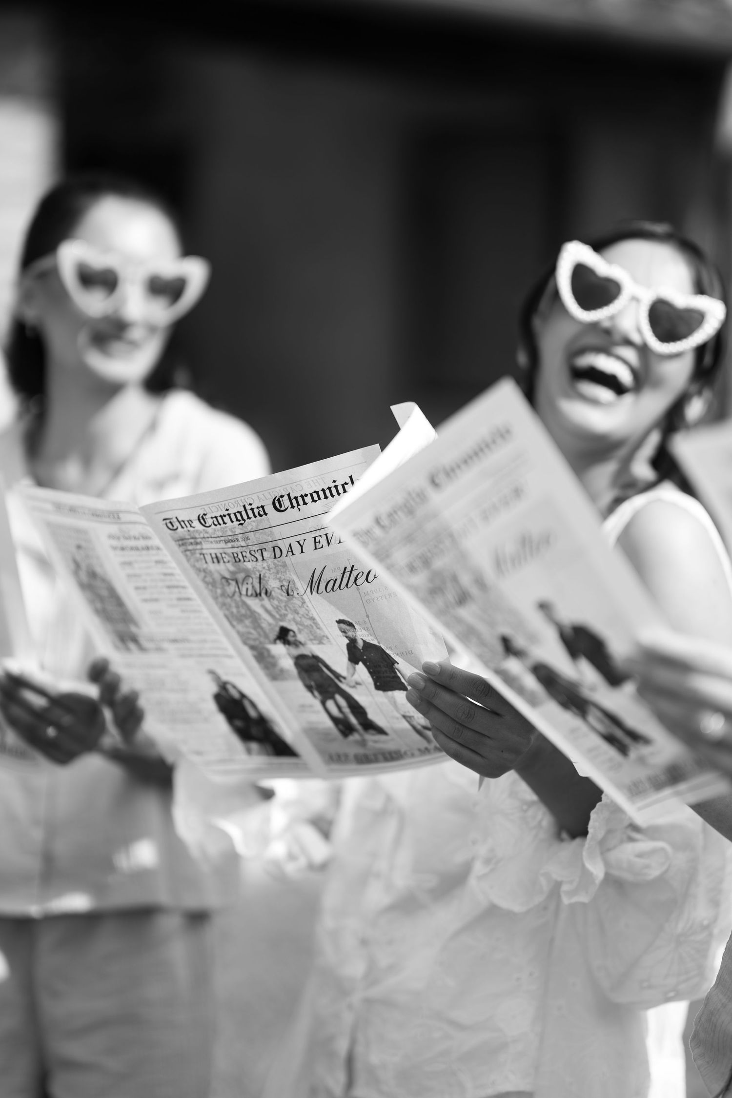 Two laughing women in heart-shaped sunglasses hold wedding-style newspapers titled The Carigila Chronicle in a black and white photo.