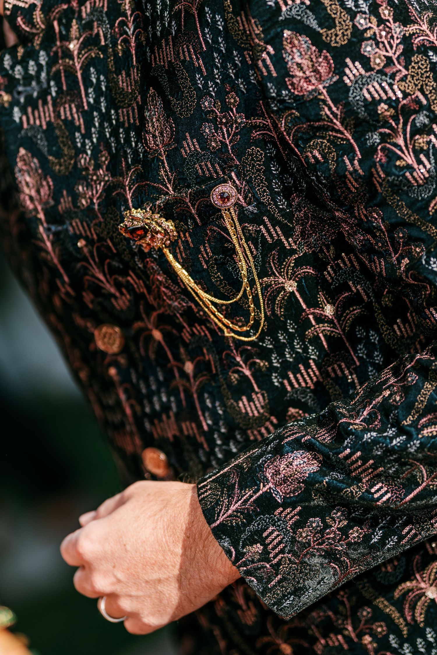 Close-up of a groom's black sherwani embroidered with pink floral threadwork and a gold chain brooch with a ruby-red stone.