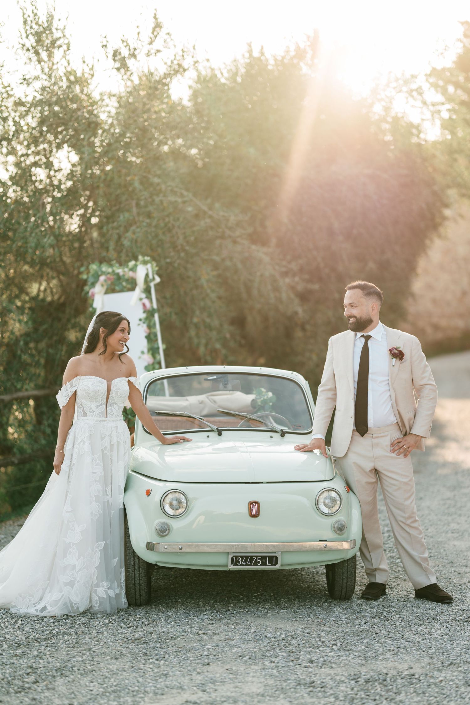 Bride in an off-shoulder lace gown and groom in a beige suit stand beside a vintage mint green Fiat 500 on a sunlit gravel road.