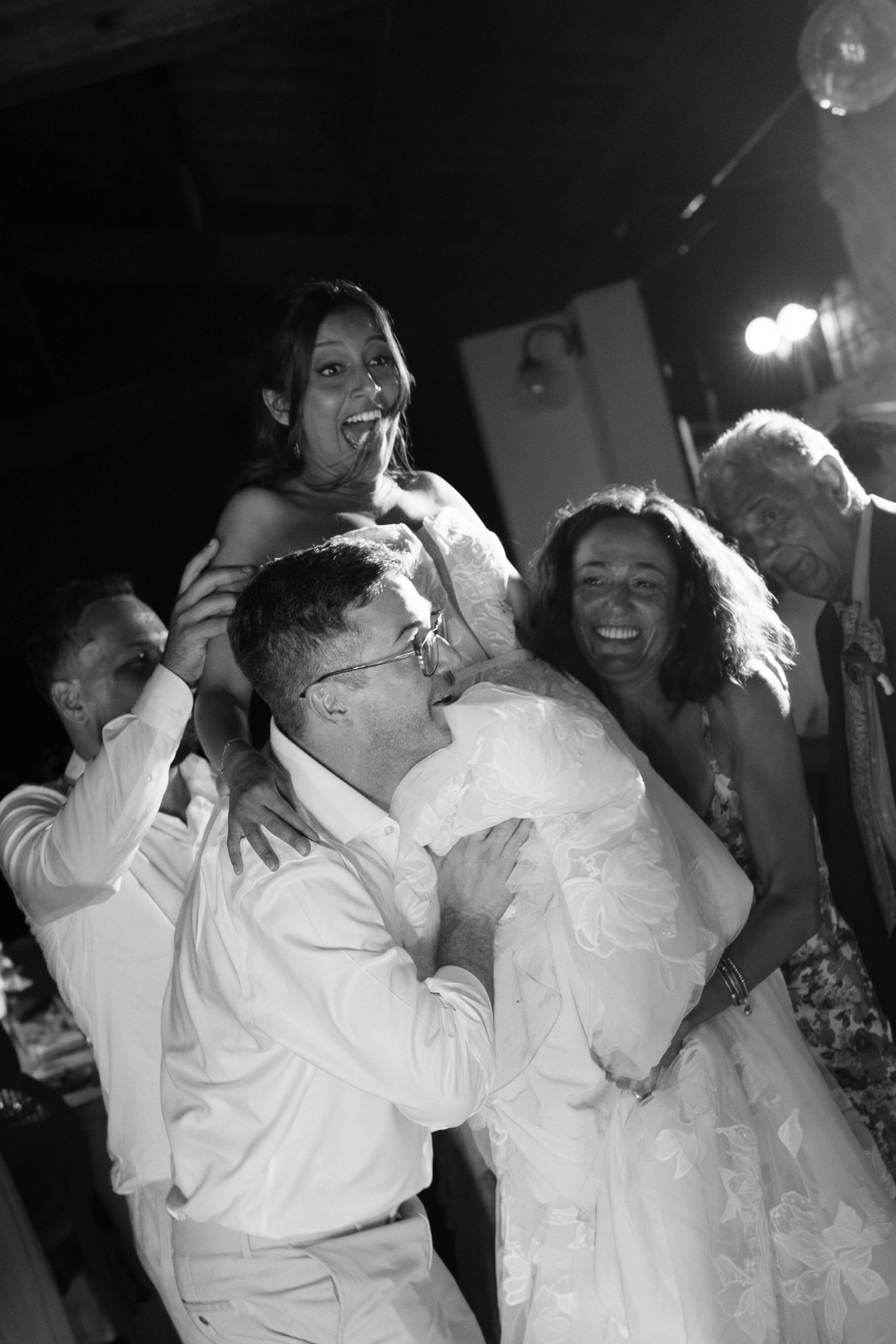 Smiling bride in a lace wedding gown is lifted by guests in a joyful black and white reception dance moment.