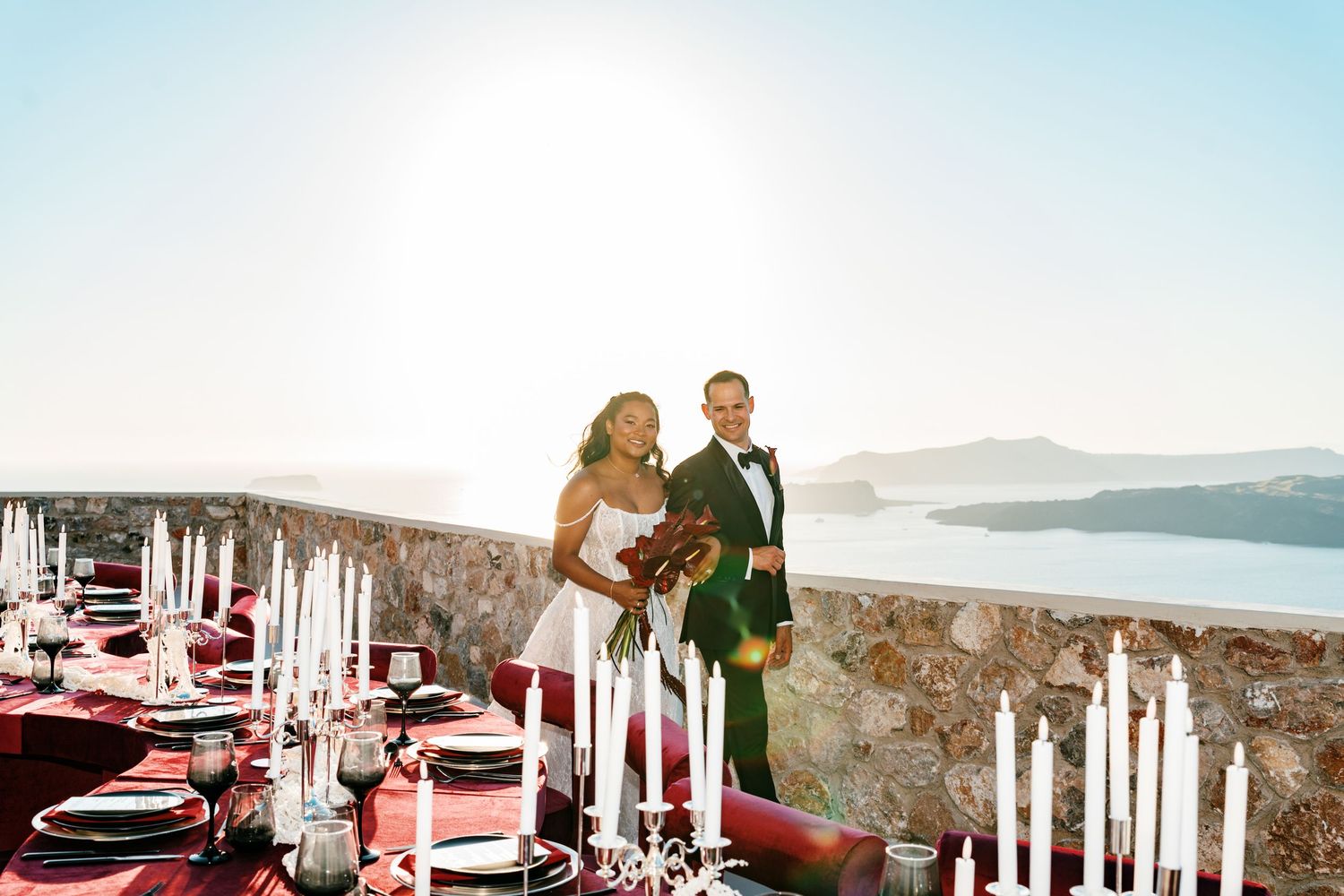 Couple poses at elegant Santorini wedding reception with red tablescape, candles, and stunning caldera ocean view backdrop.