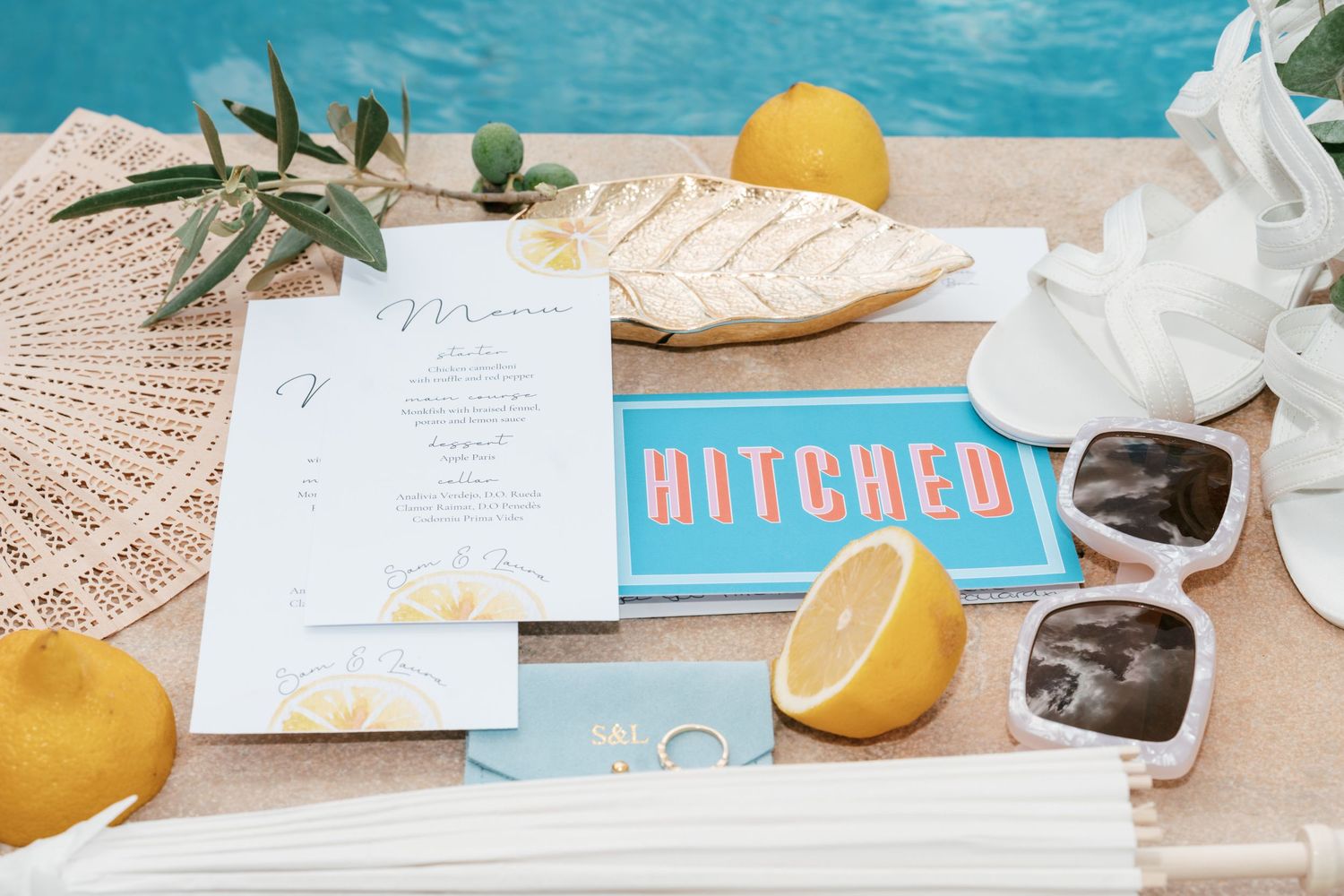 Colorful wedding stationery flat lay with lemons, sunglasses, and a blue HITCHED sign by a pool. Mallorca wedding filmmaker, videographer
