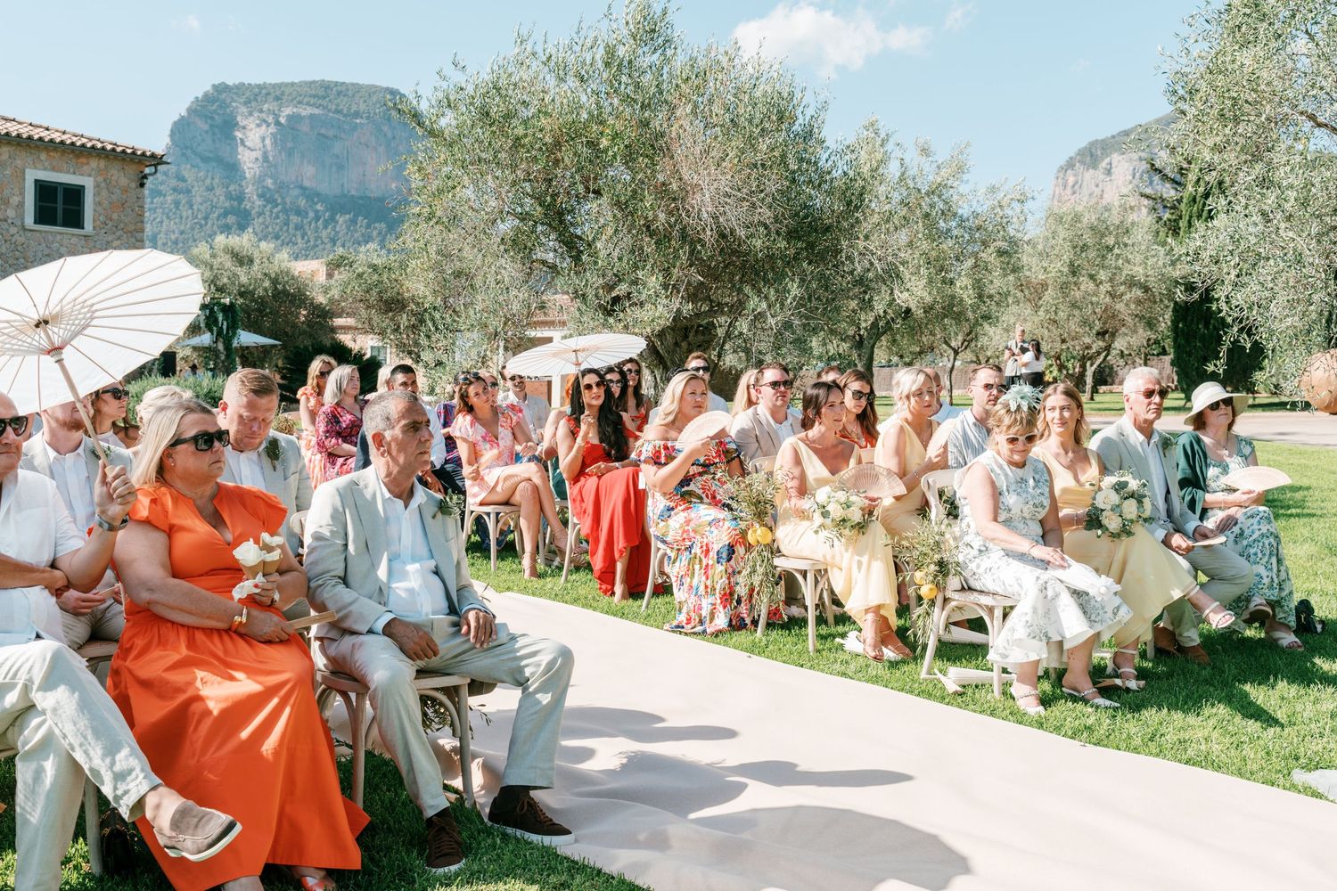 Guests seated outdoors at a sunny wedding ceremony, surrounded by olive trees and rocky hills in the background. Mallorca wedding photograph