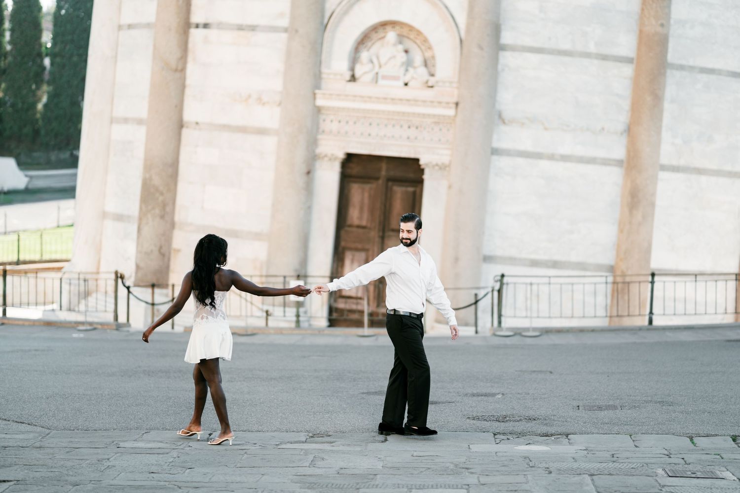 Couple holding hands and dancing playfully on a stone plaza in front of a historic white marble building with a wooden door.