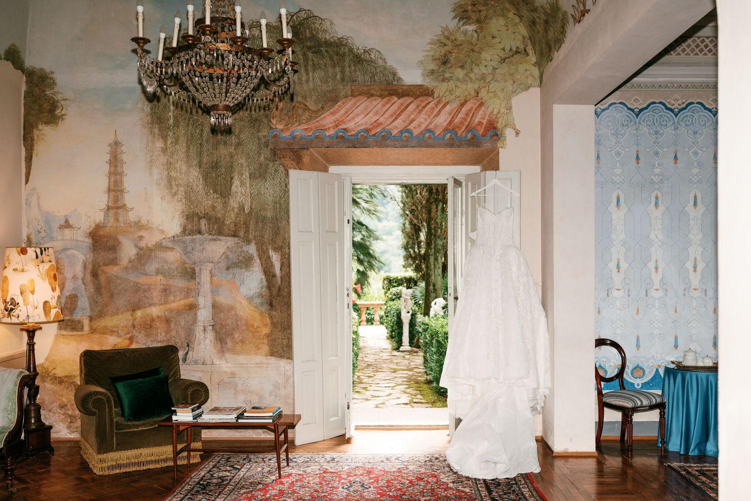 A white wedding gown hangs in an open doorway of an elegant room with a crystal chandelier, scenic mural walls, and a garden view beyond.