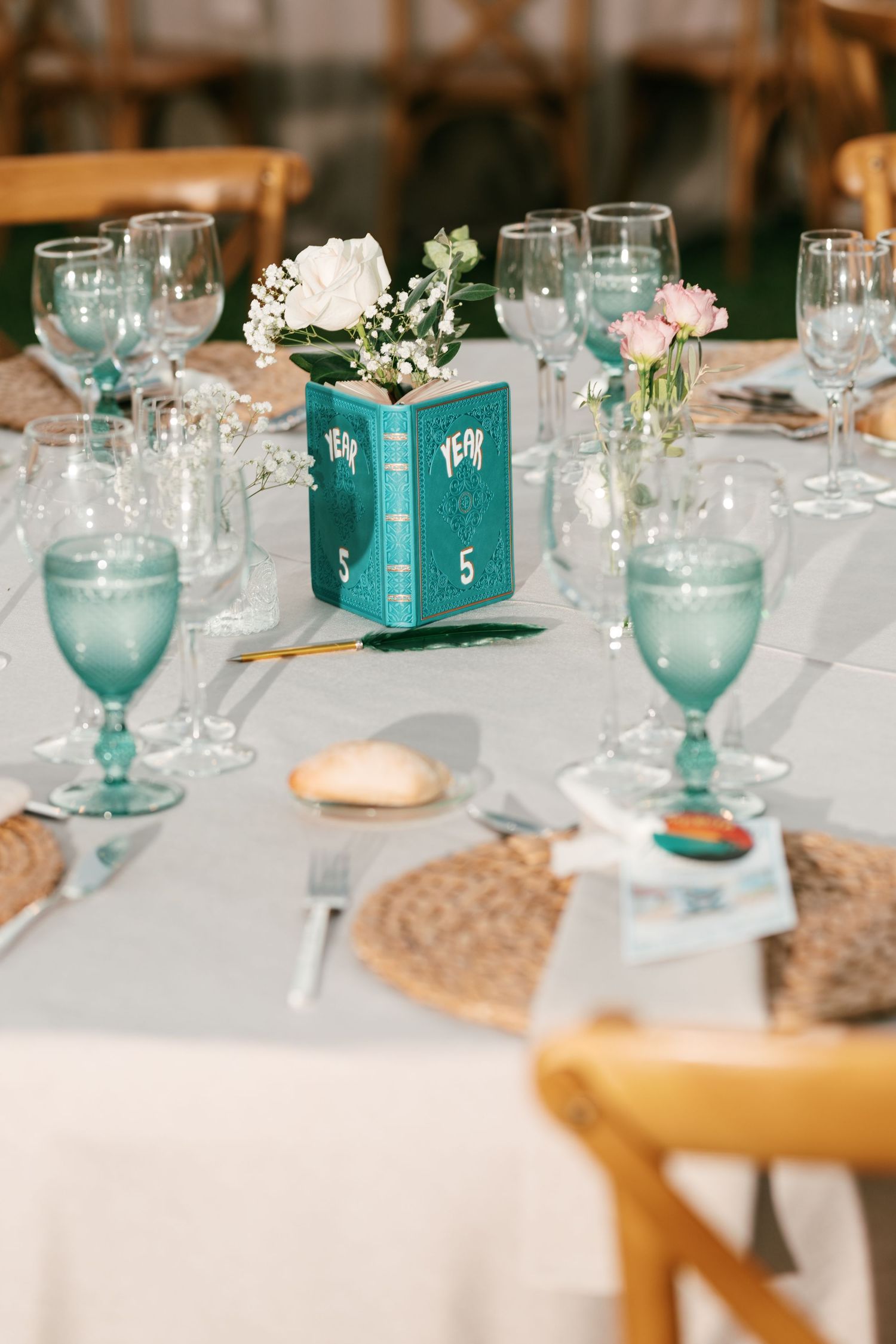 Wedding reception table with a teal book vase holding white and pink flowers, green goblets, woven placemats and wooden chairs.