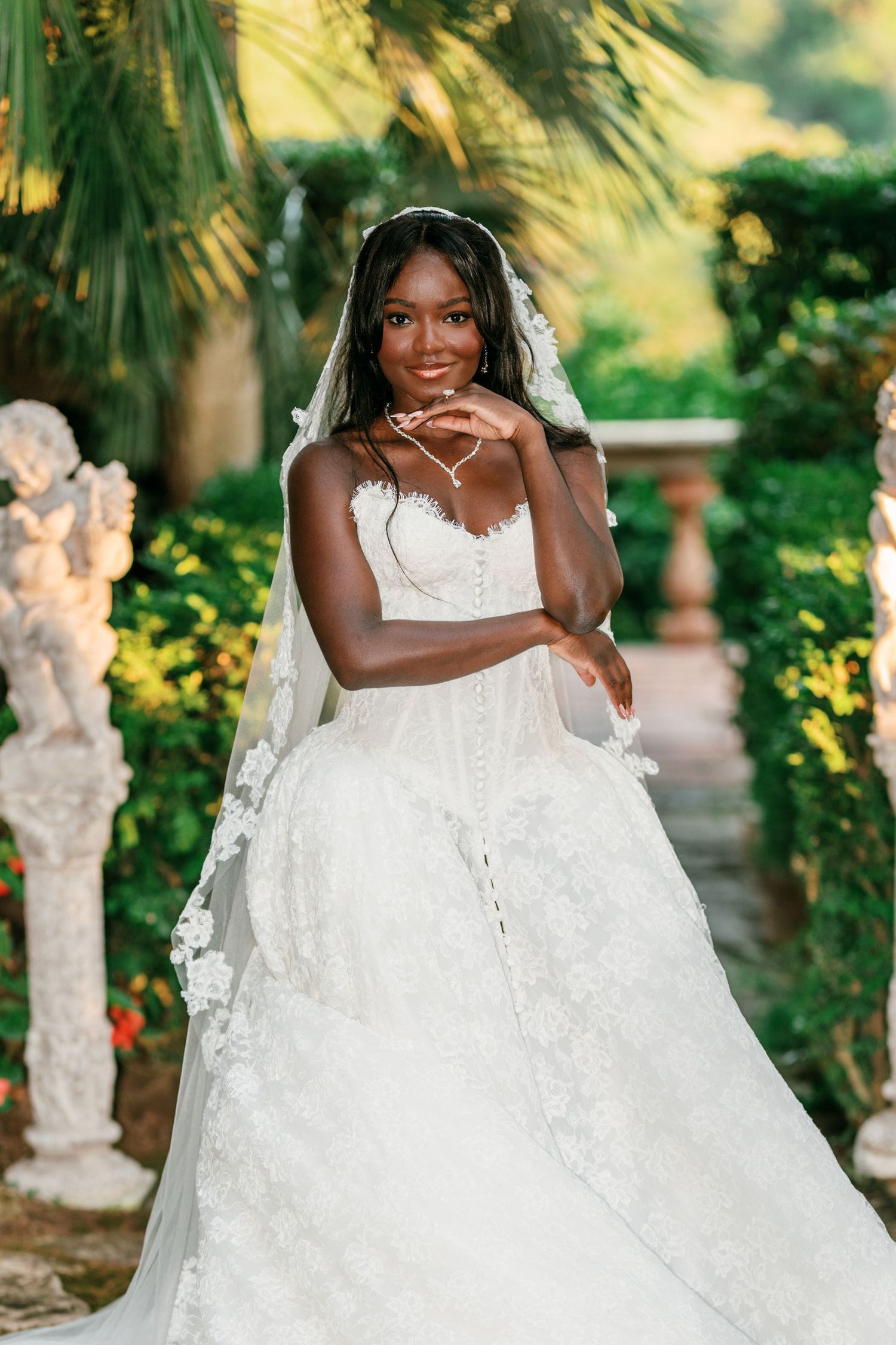 Smiling bride in a strapless white lace wedding gown and veil poses in a lush garden with stone cherub statues.