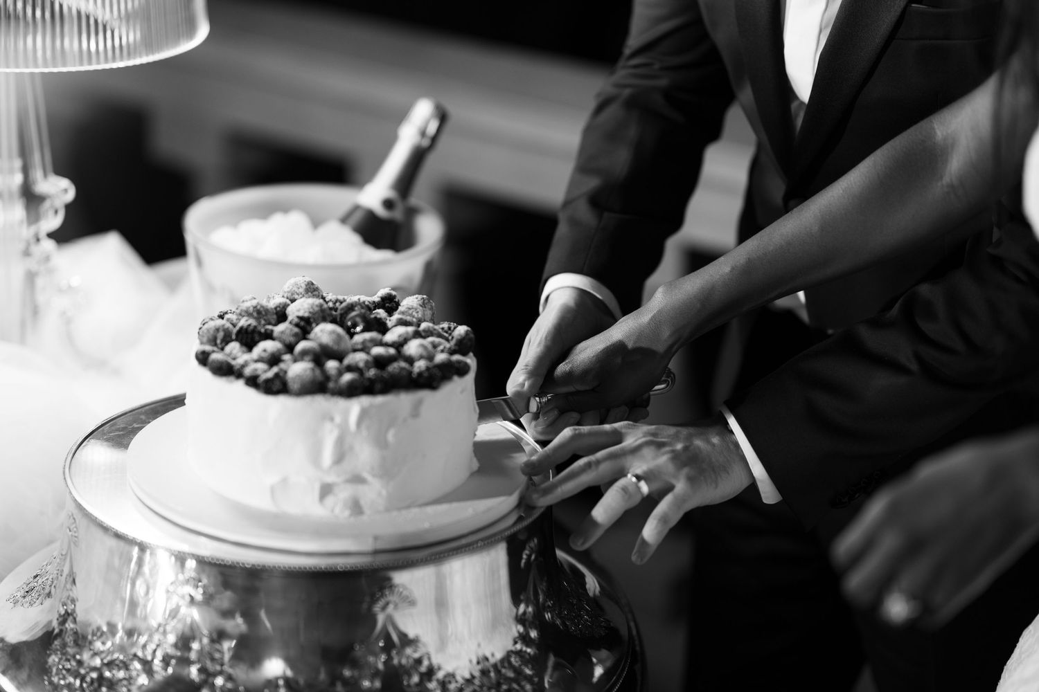Black and white close-up of a bride and groom cutting a berry-topped wedding cake together with a champagne bottle behind.