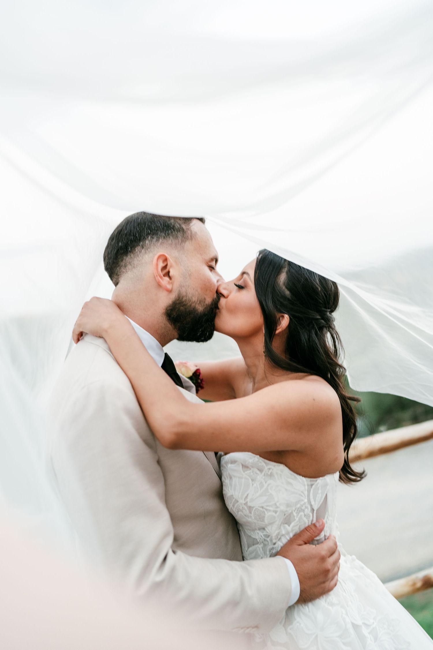 Bride in a lace wedding gown and groom in a beige suit share a kiss beneath a flowing sheer veil outdoors.
