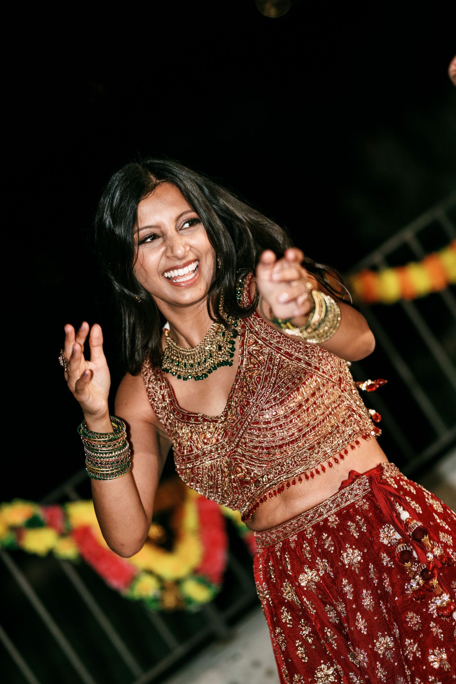 Smiling woman in a red and gold embroidered lehenga with layered green necklace and bangles dancing joyfully at a festive celebration.