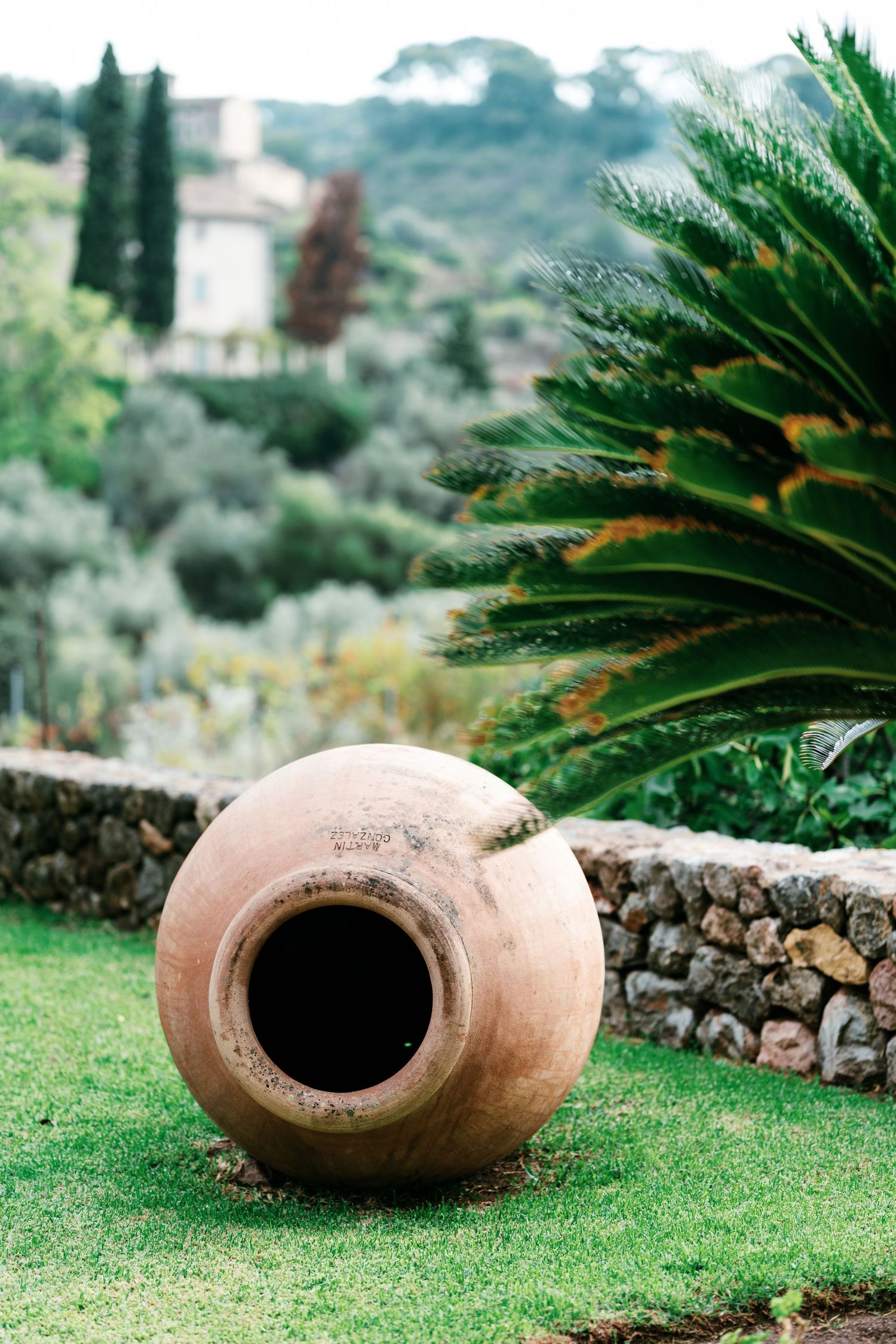 Large terracotta amphora resting on a green lawn beside a stone wall, with a palm, cypress trees and a villa in the background.