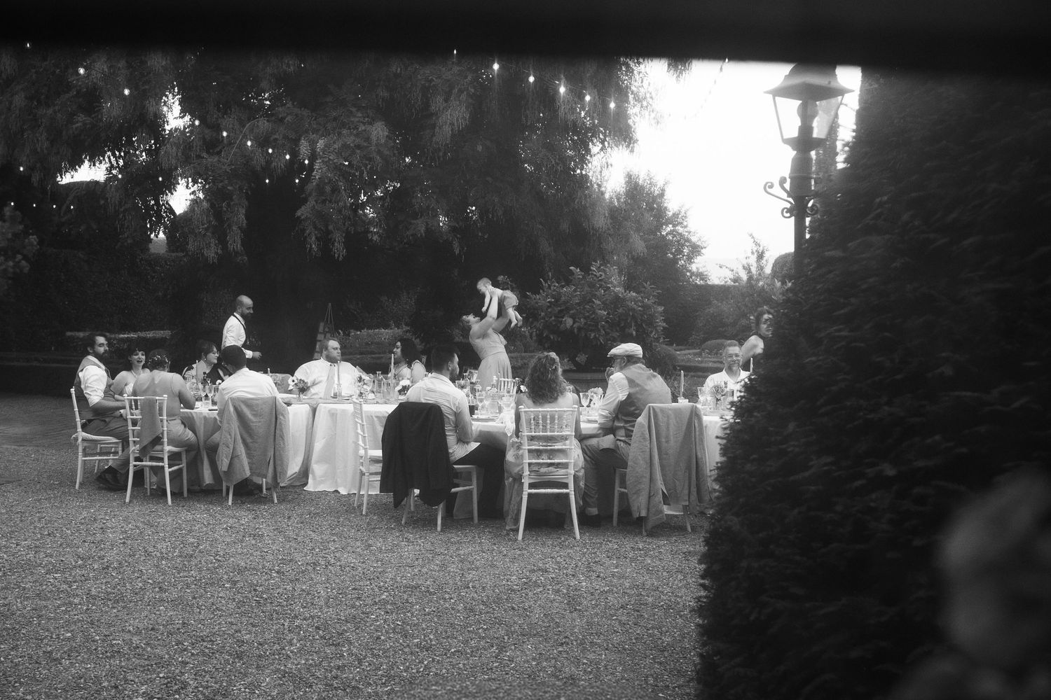 Black and white photo of guests seated around a long table at an outdoor garden dinner party under tall trees, with one person standing.