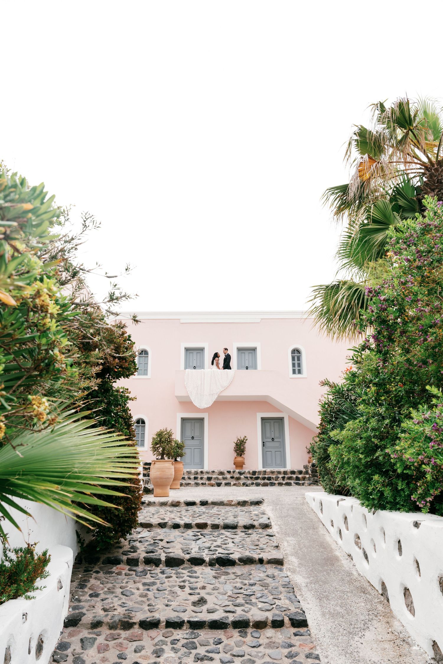 Couple on balcony of pink Mediterranean villa with stone steps, lush greenery, and palm trees in Santorini, Greece.