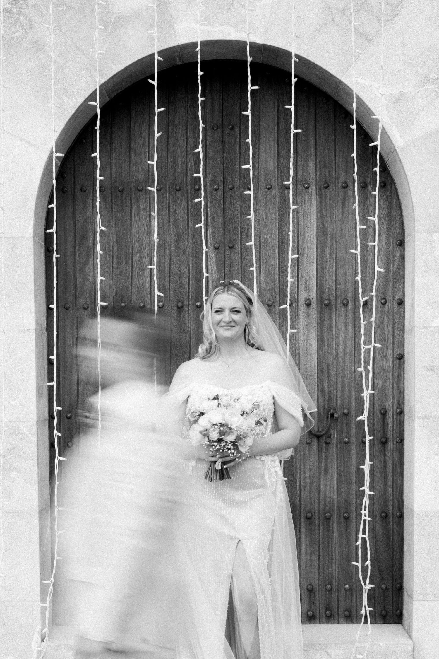 Bride in white gown holding bouquet stands before arched wooden door with fairy lights in black and white photo. Mallorca wedding photo film