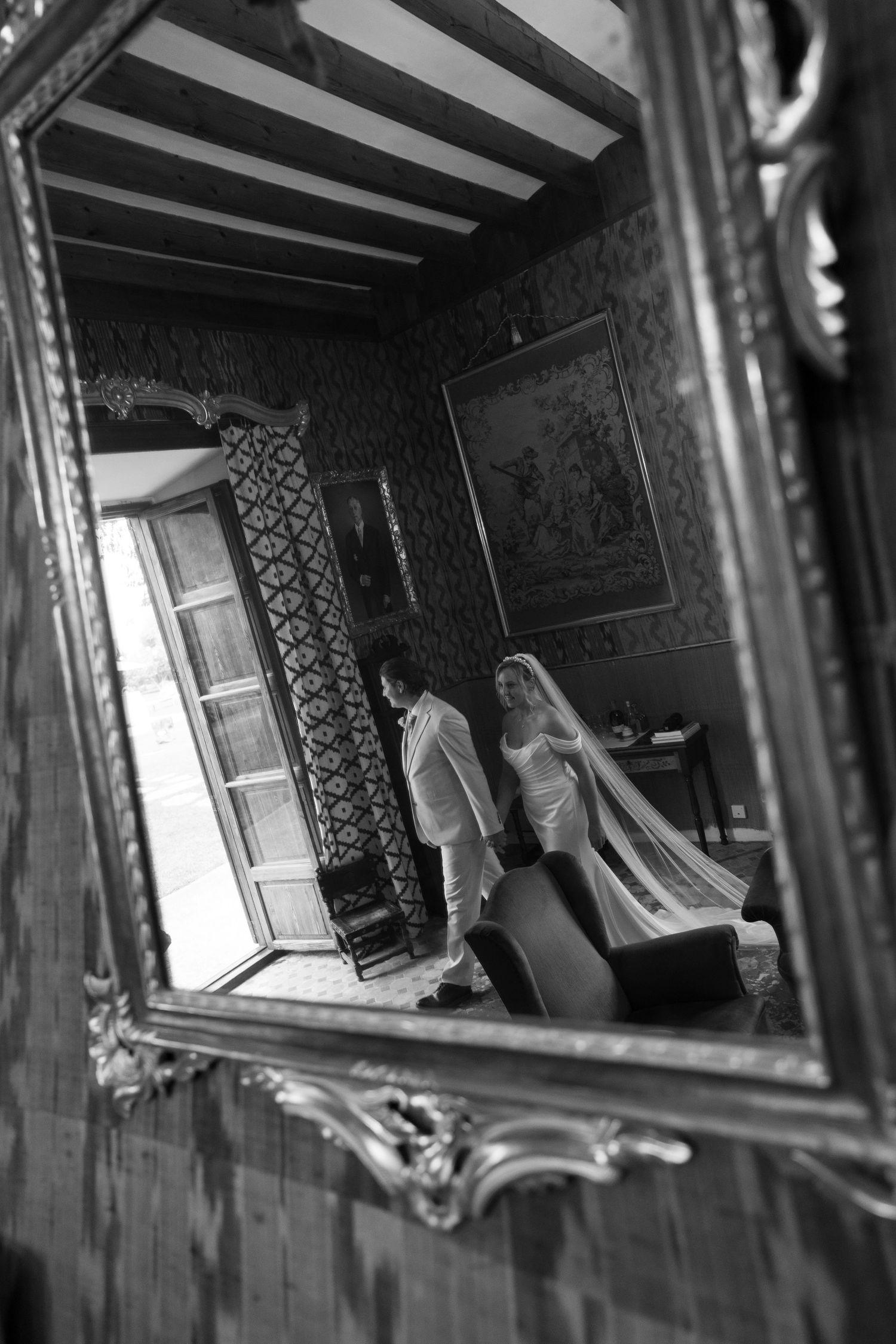 Black and white mirror reflection of a bride in a long veil and a groom in a suit walking through an ornate room with patterned walls.