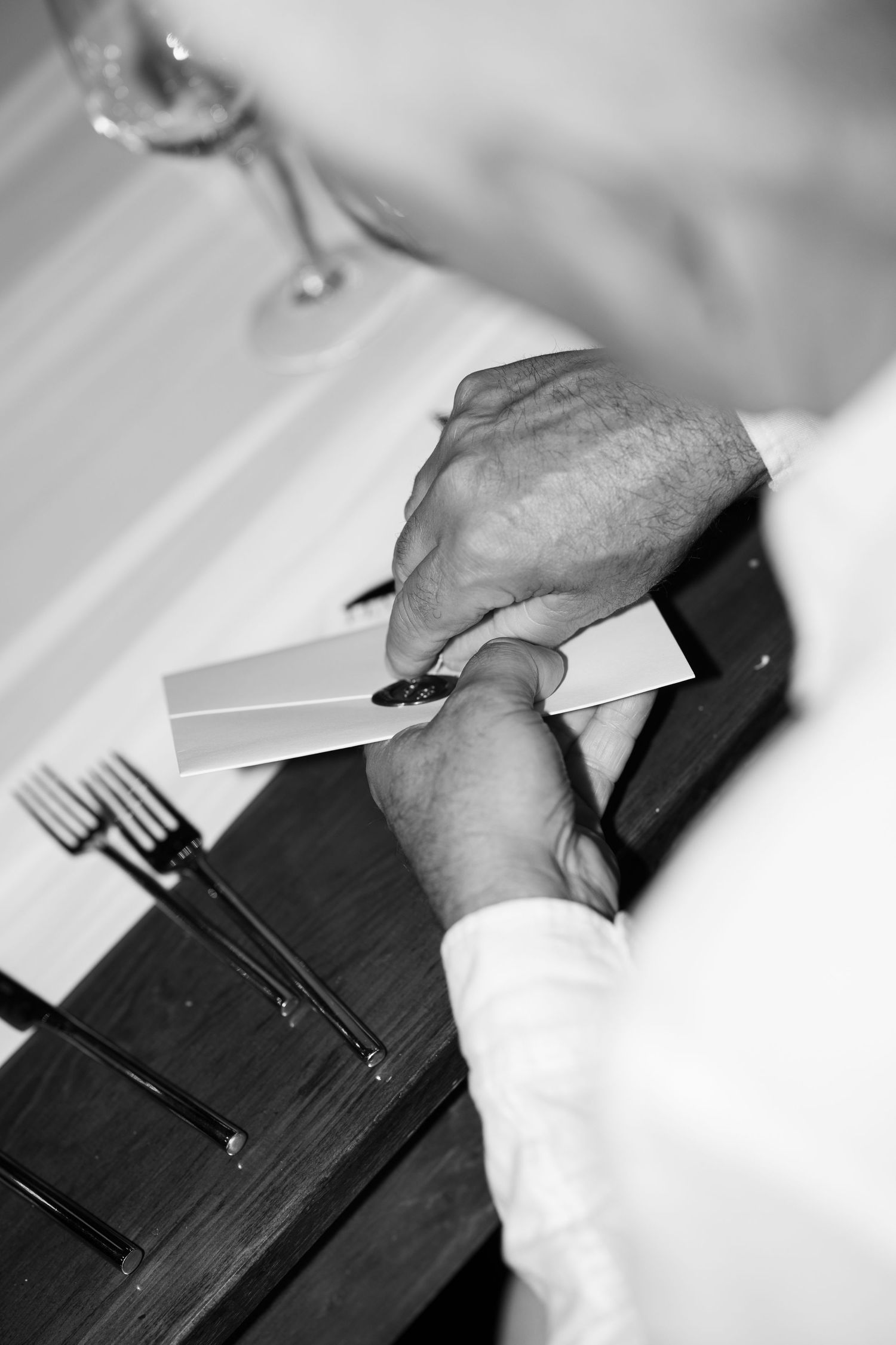 Groom in white shirt carefully reads place card at elegant dark wood wedding reception table with forks and champagne glass.