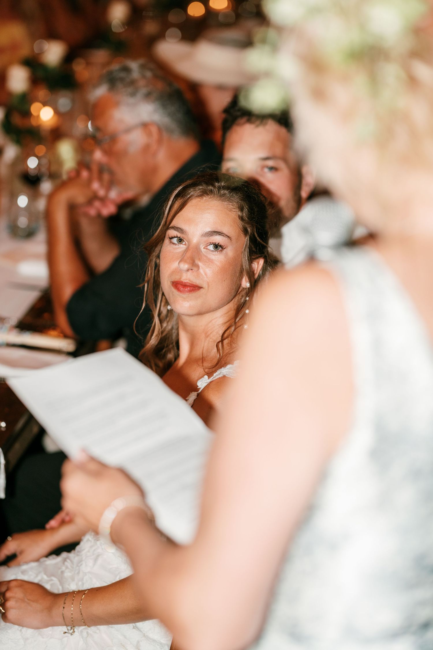 Bride listens attentively during wedding speech at elegant reception dinner, with guests seated at candlelit table.