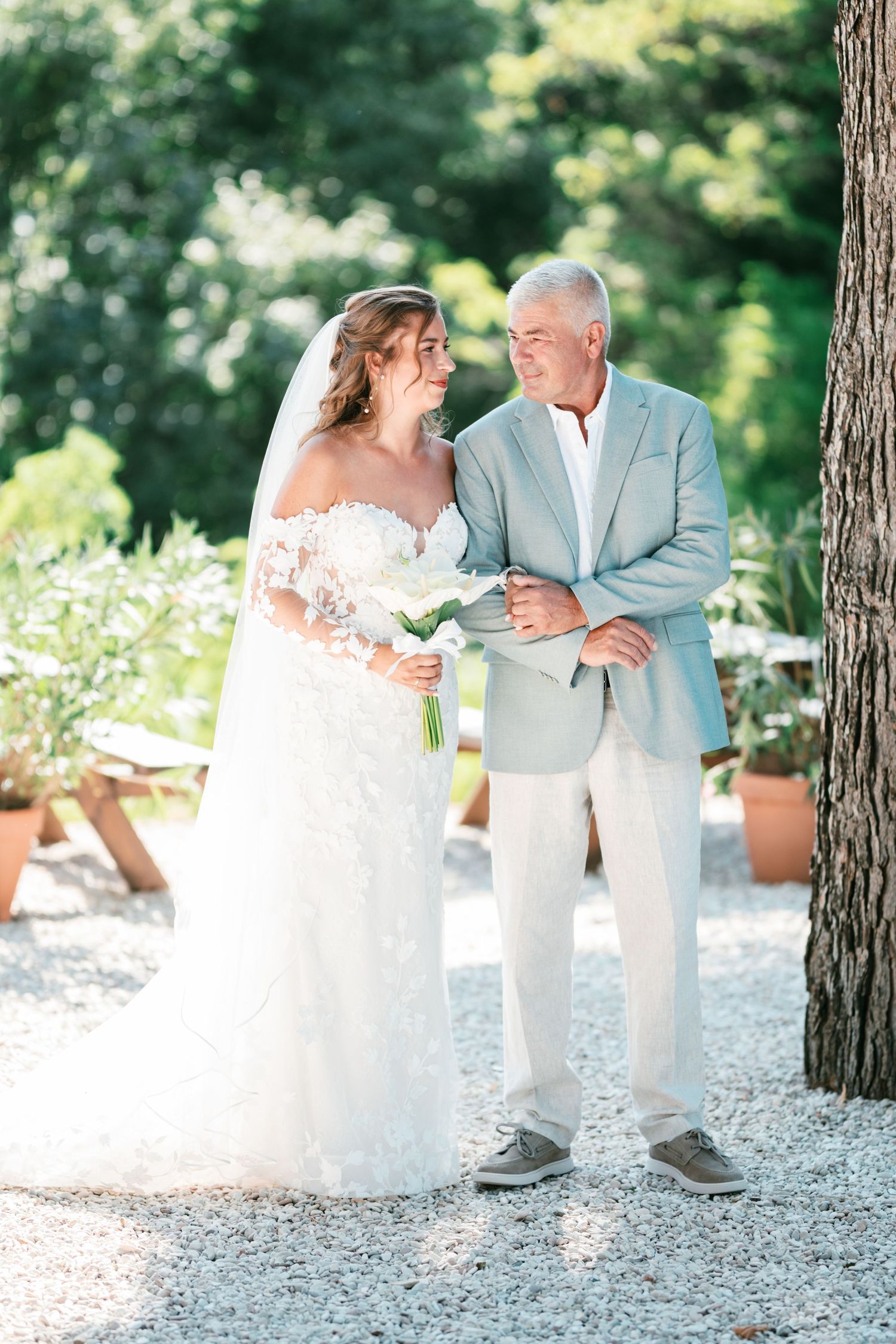 Bride in lace off-shoulder gown walks with father in light gray suit on gravel path.