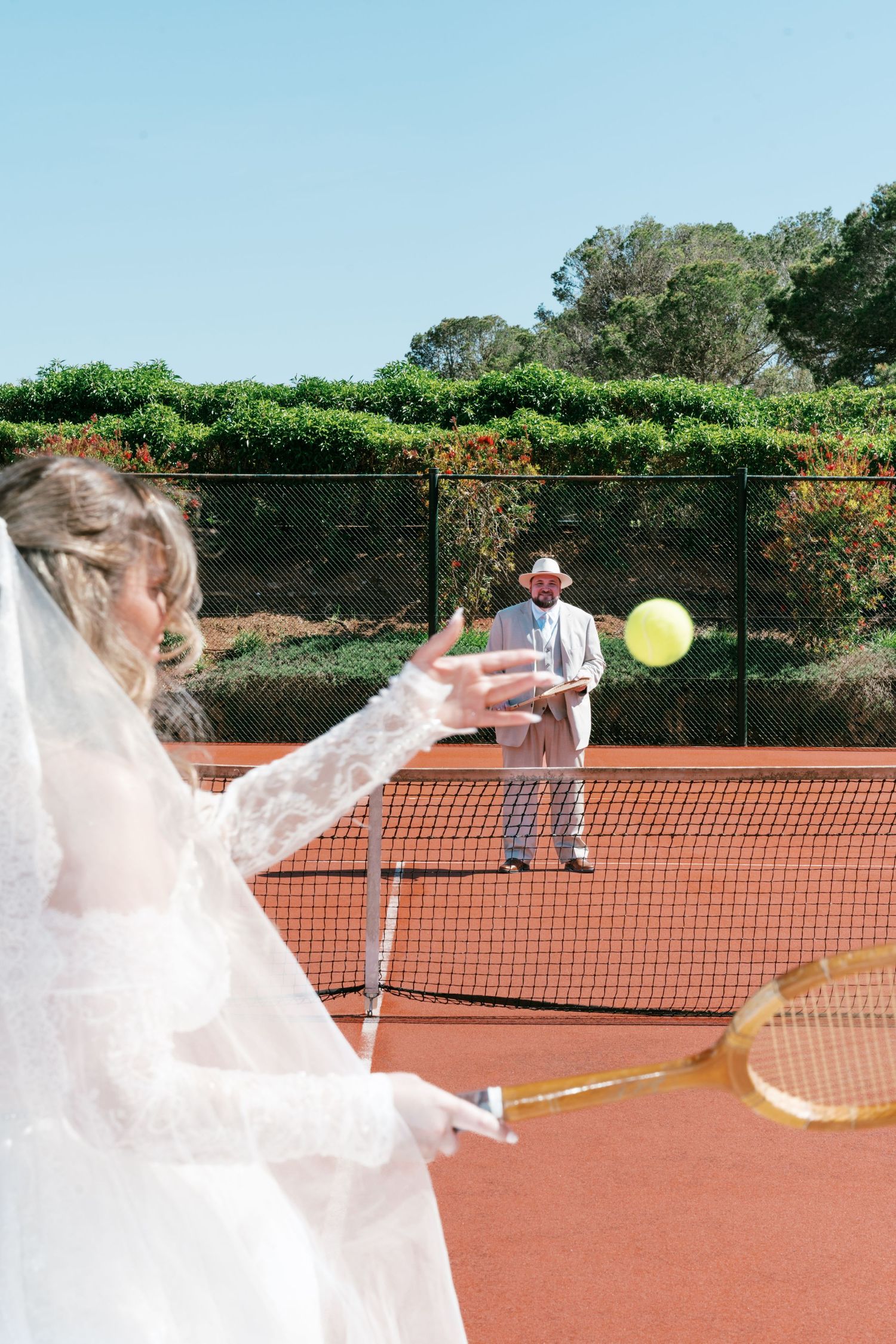 Bride in white wedding dress playing tennis on clay court with groom in suit and hat across the net on sunny day in Mallorca.