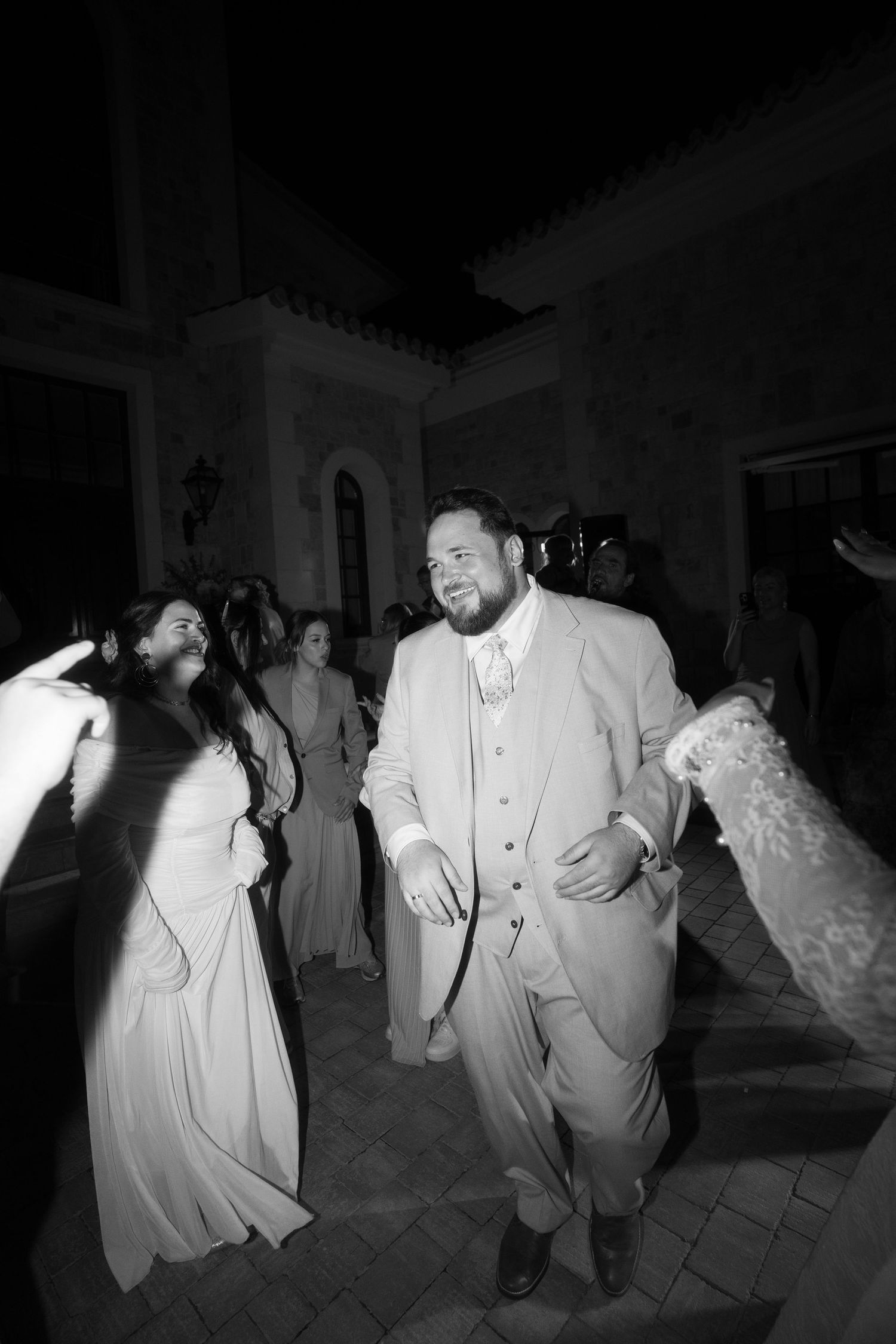Black and white photo of a groom in a suit dancing joyfully with guests at an outdoor wedding reception at night in Mallorca.