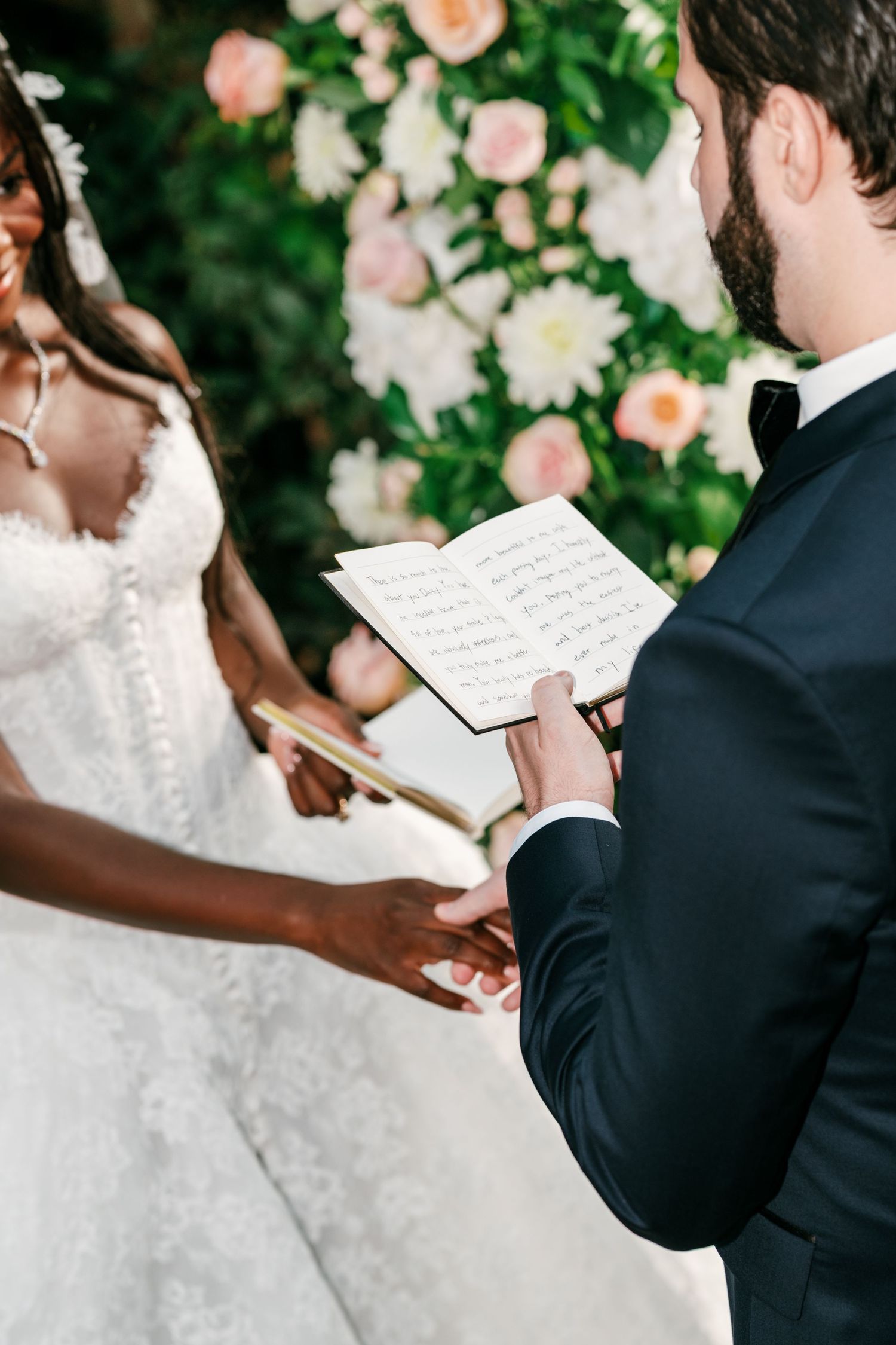 Groom in a navy suit reads handwritten vows from a notebook while holding hands with the bride in a lace gown near pink floral arrangements.