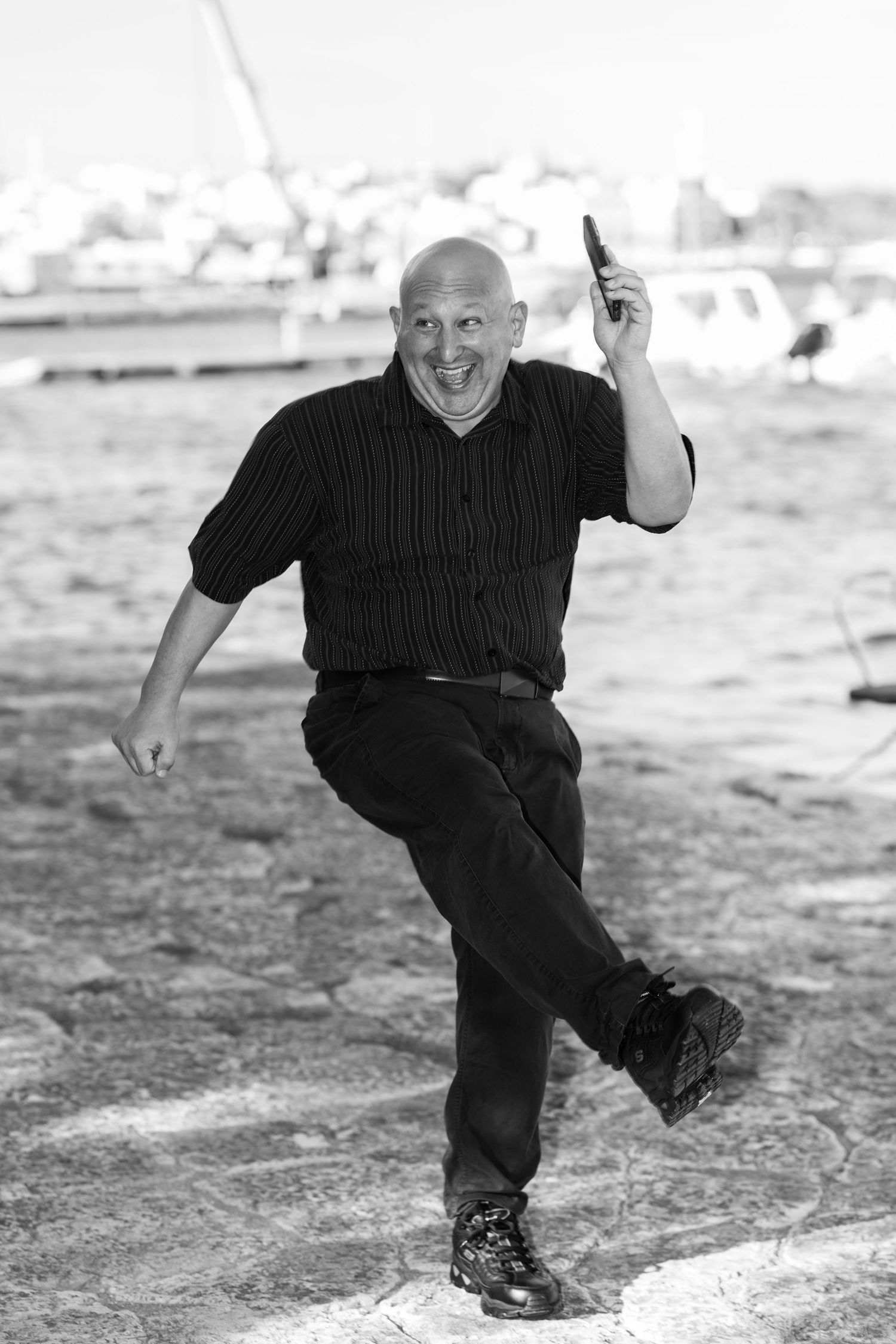 A smiling bald man in a dark striped shirt lifts one knee and holds a phone to his ear while dancing playfully outdoors near a harbor.