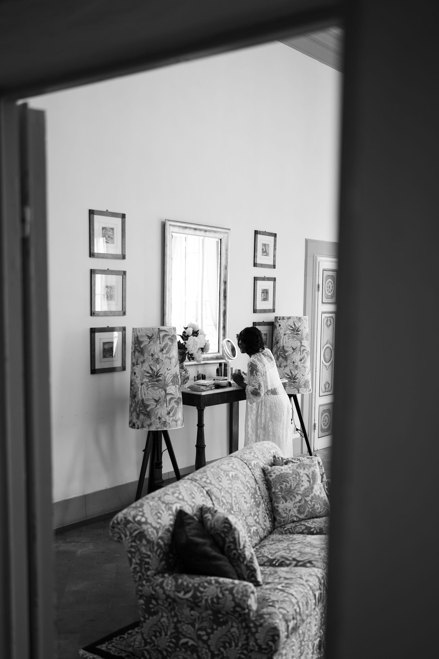 Black and white photo of a woman in a robe at a vanity table in an elegant room with a floral sofa, framed art, and patterned lamps.