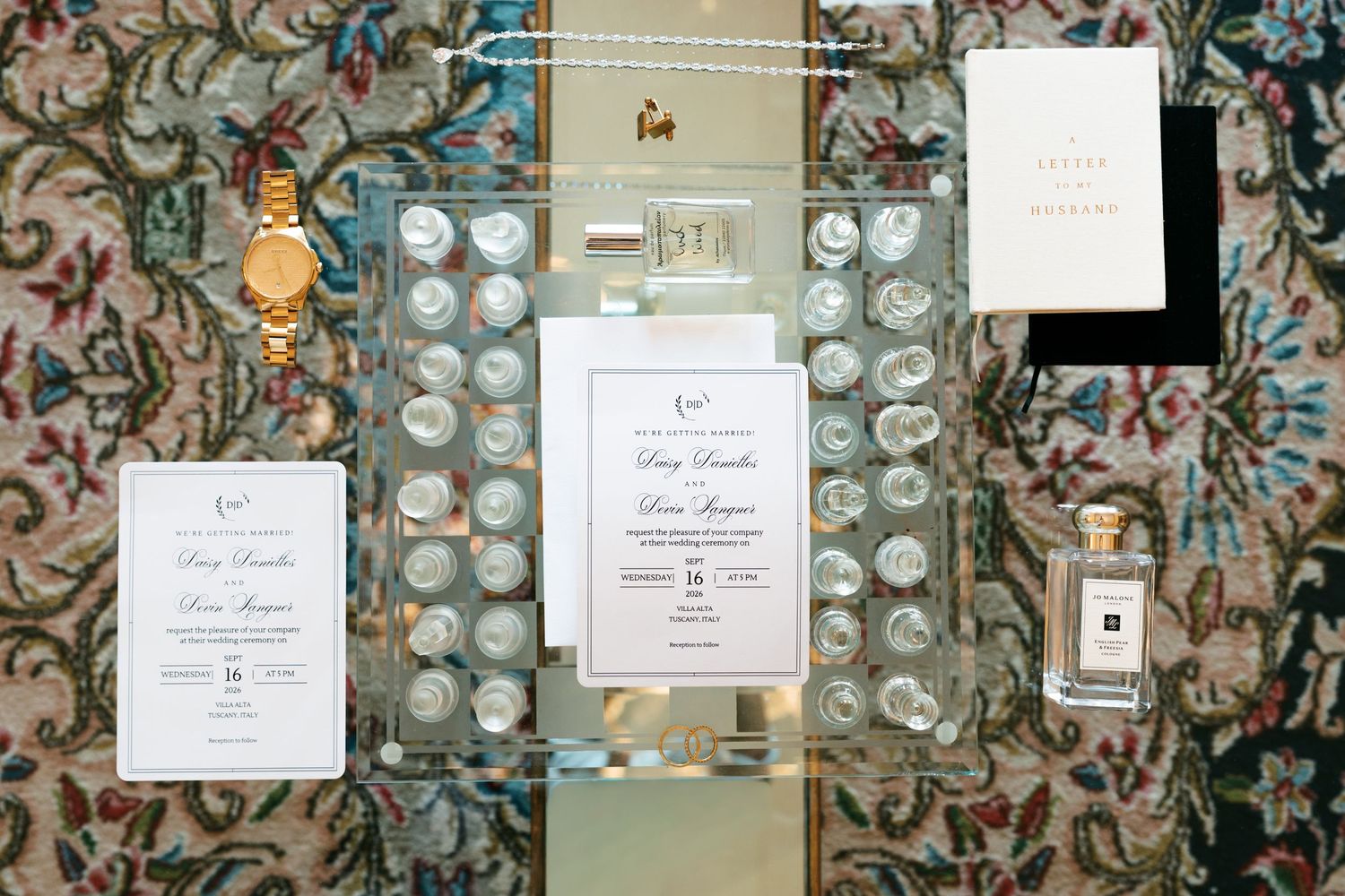 Flat lay of wedding invitation suite on a clear acrylic tray with a gold watch and cologne bottle atop an ornate patterned rug.