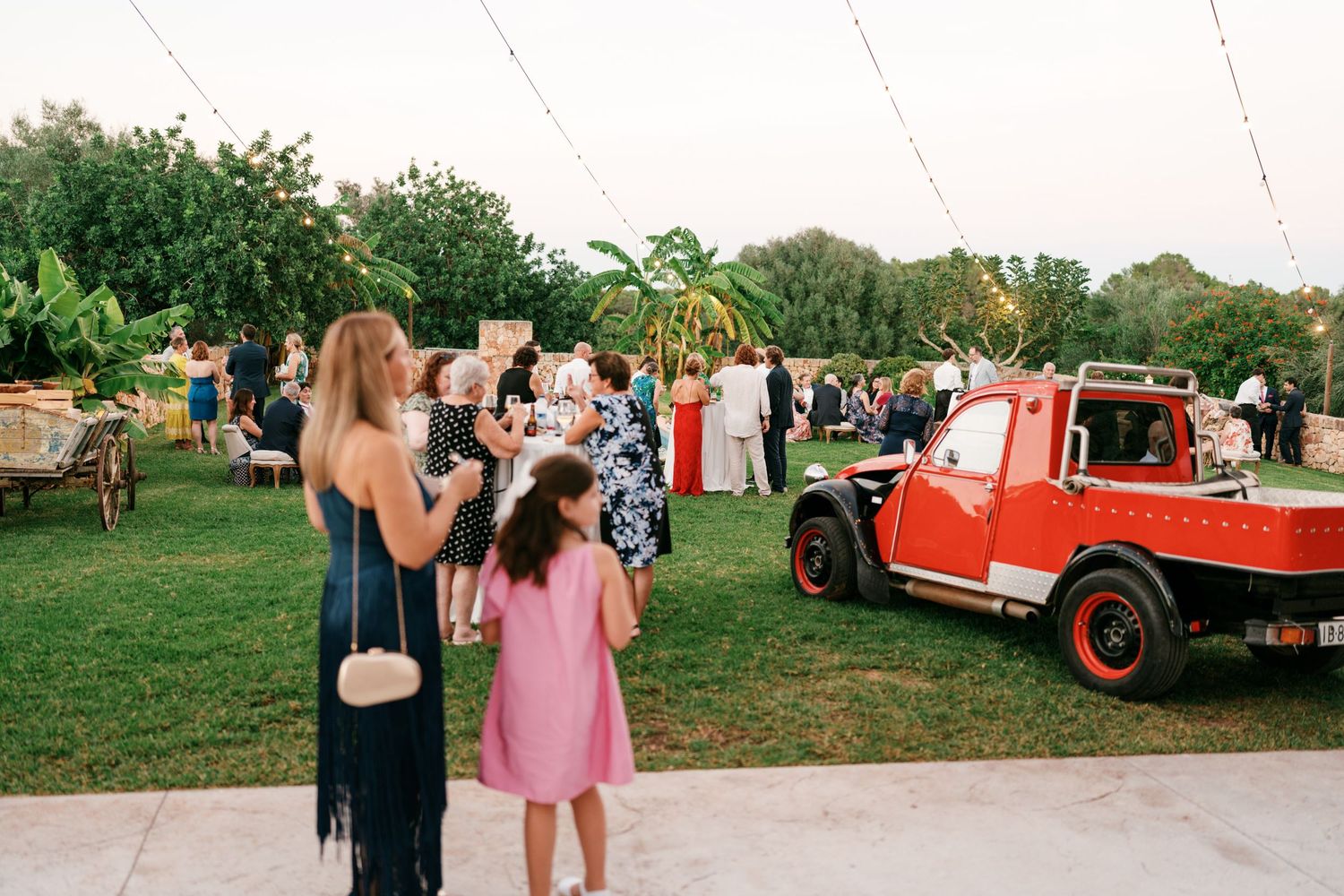 Wedding guests mingle on a lawn near a vintage red pickup truck as a woman and girl in a pink dress look on at an outdoor reception.