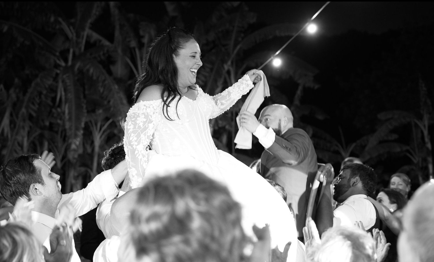 Smiling bride in a white lace gown is lifted high on a chair by cheering wedding guests during a joyful outdoor evening reception.
