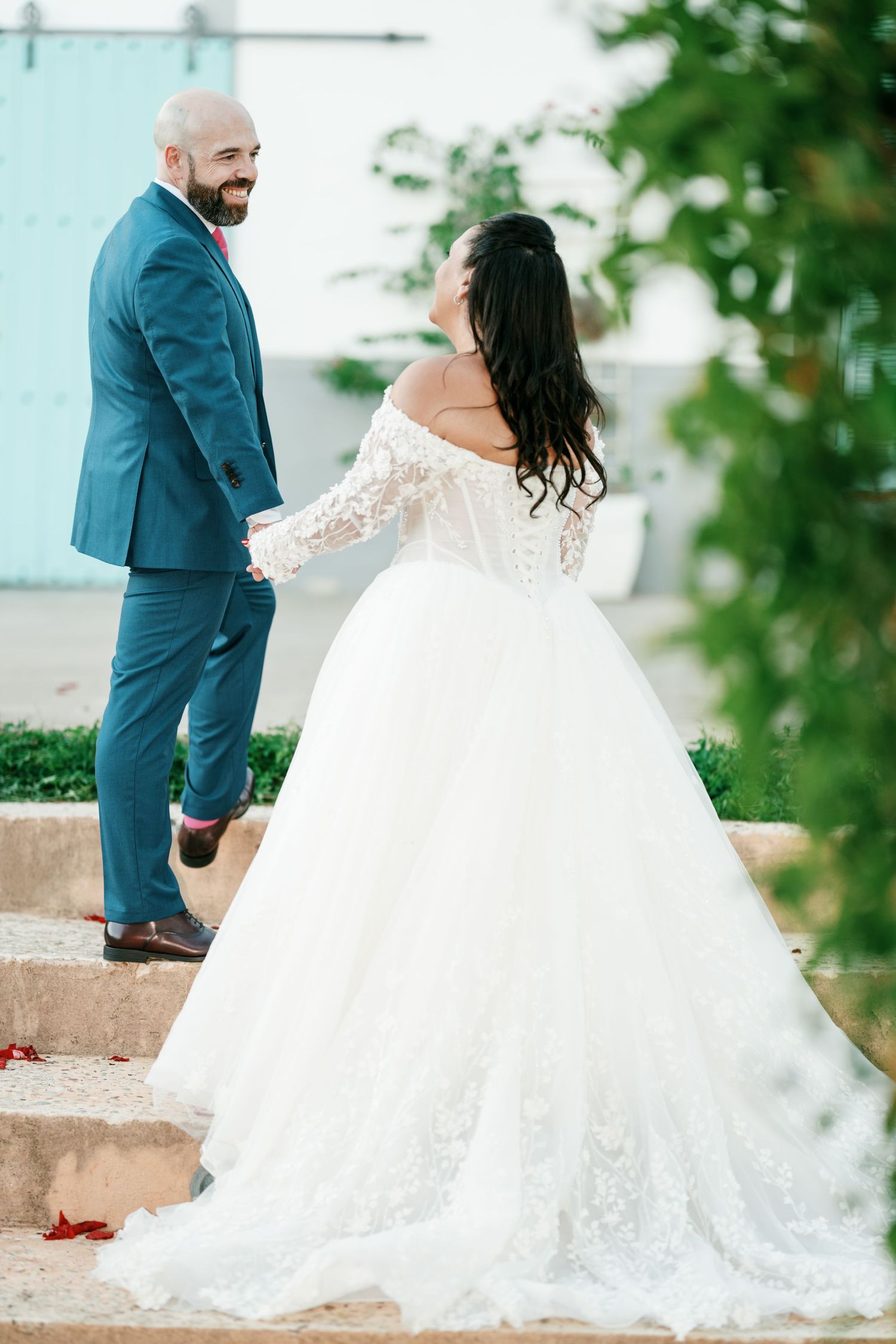 Groom in a teal suit holds hands with a bride in a lace off-the-shoulder ball gown as they walk up outdoor stone steps.