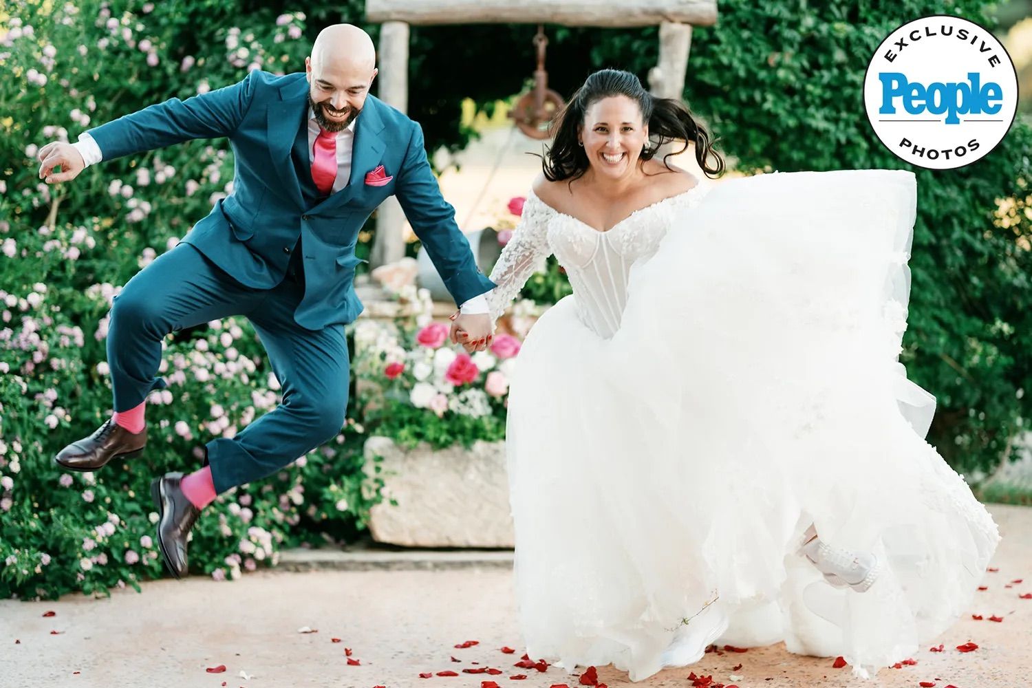Joyful bride in a white off-the-shoulder gown and groom in a blue suit with pink socks leap and laugh among rose petals in a garden.