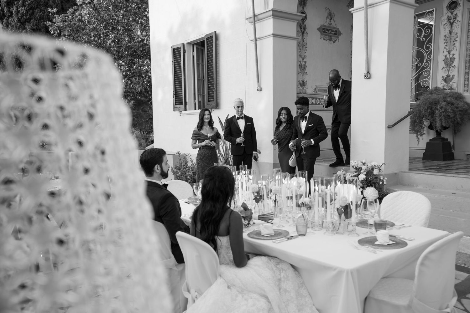 Wedding guests in formal attire stand near an elegant outdoor reception table set with flowers and candles as a couple sits together.