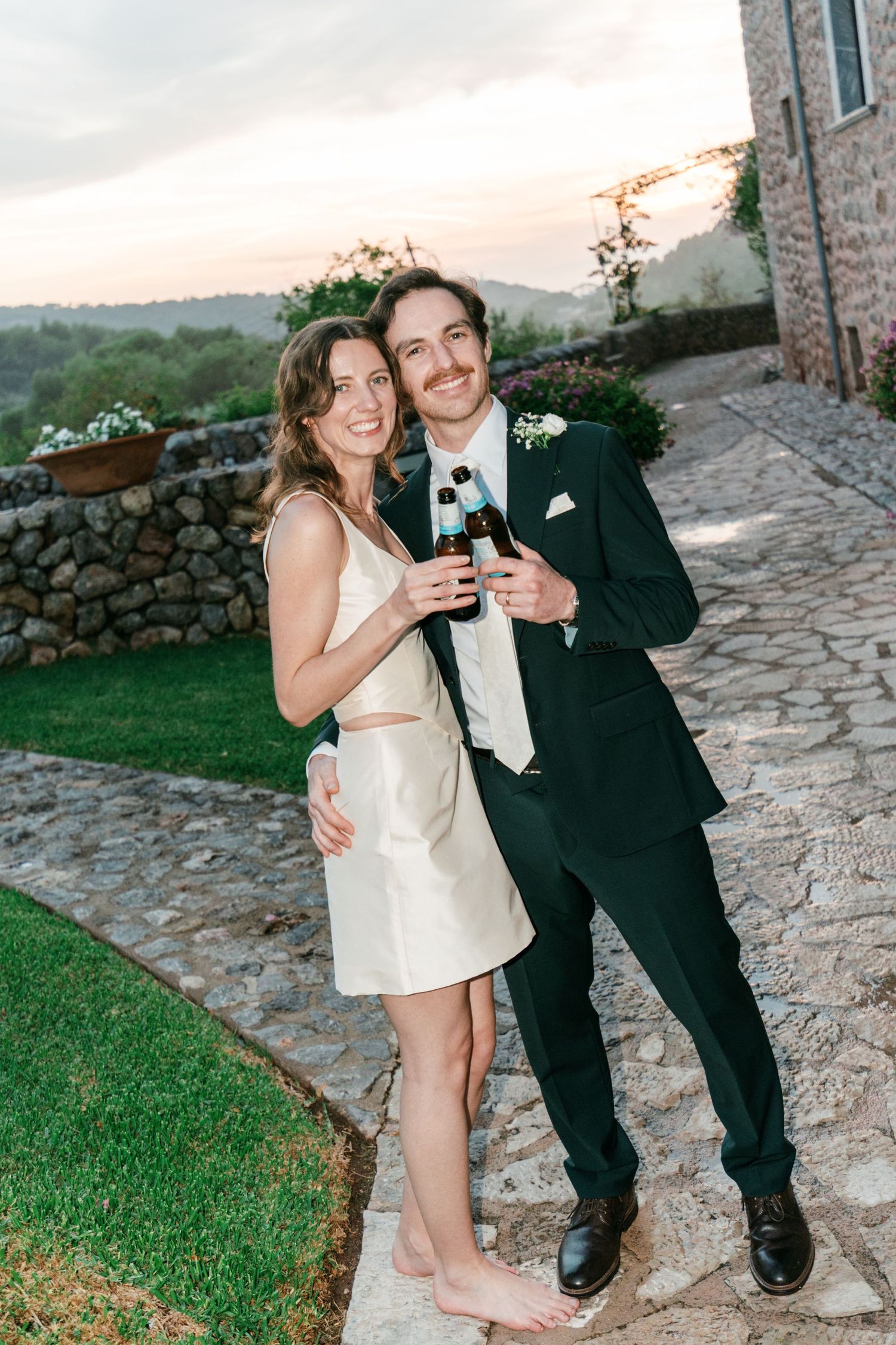 A smiling couple in a green suit and cream dress hold champagne bottles on a stone path at a countryside wedding.