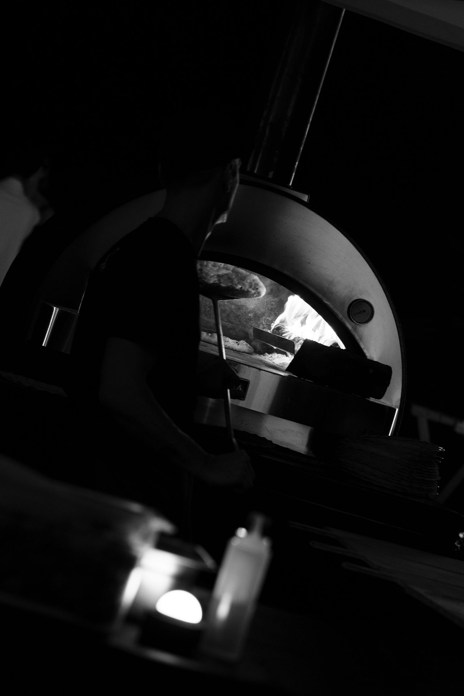 Black and white photo of a chef in silhouette using a long peel to bake pizza in a glowing wood-fired oven.
