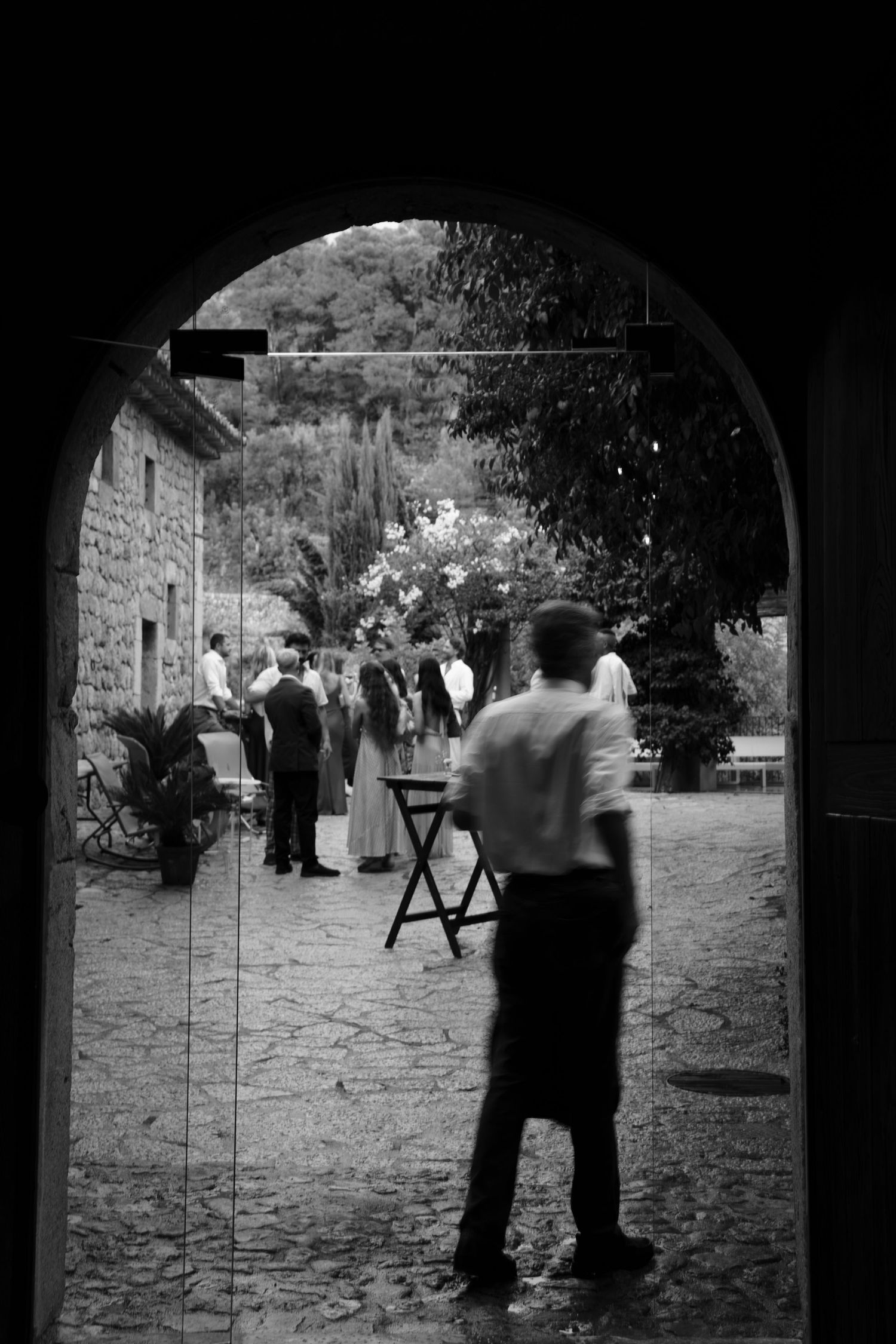 Black and white view through an arched doorway of a waiter and wedding guests mingling in a stone courtyard with trees.