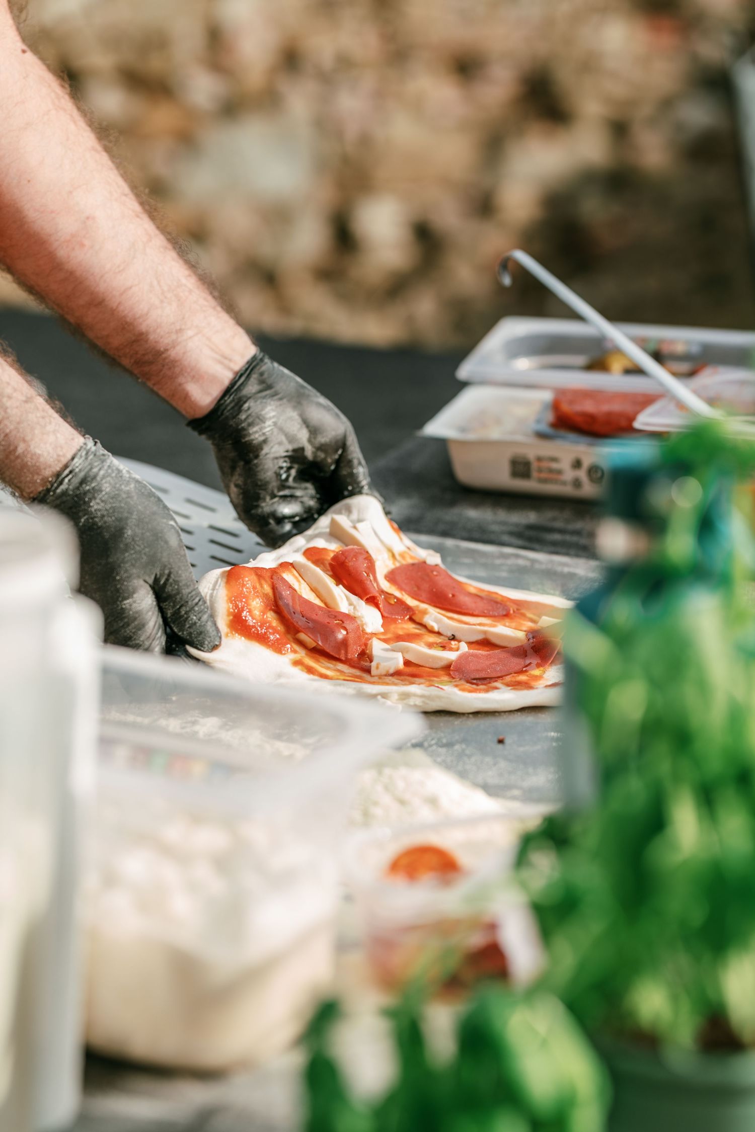 Gloved chef hands assembling a fresh pizza with tomato sauce and cheese on a rustic outdoor prep table with herbs and ingredient containers.