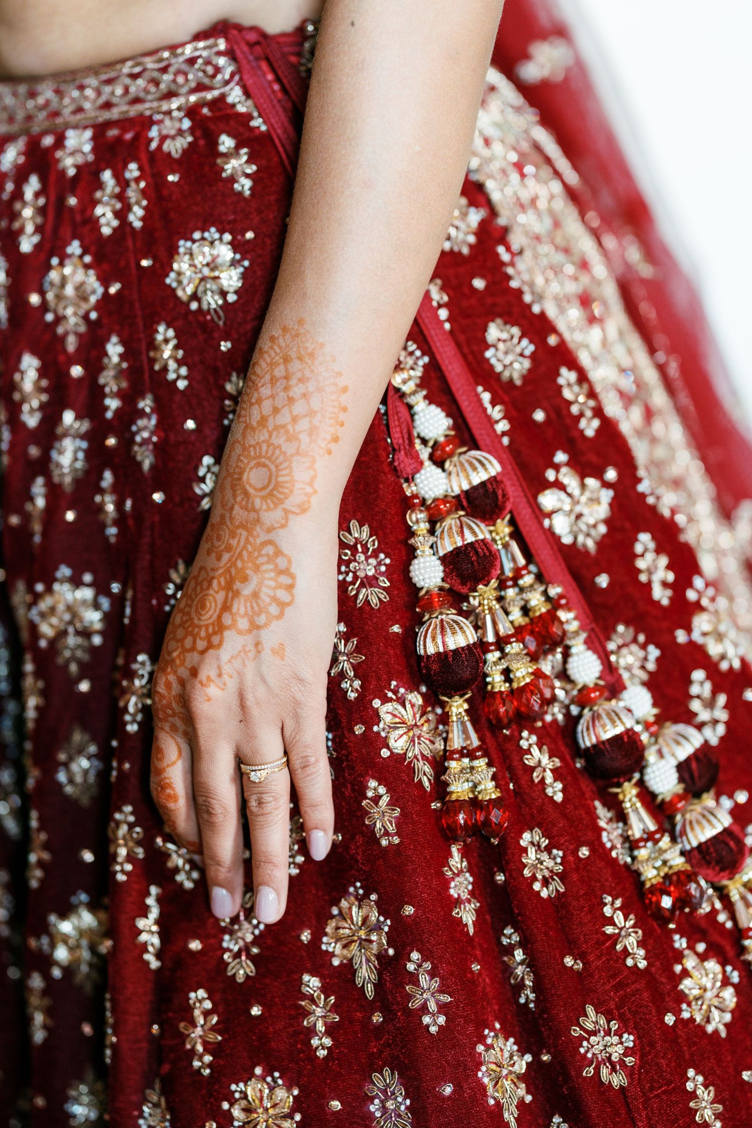 Bride's hand with delicate orange henna design resting on a red embroidered lehenga with ornate tassels and a gold ring.