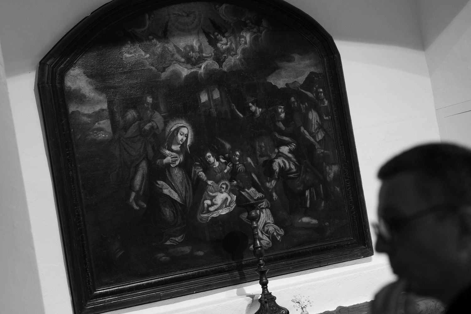 Black-and-white photo of a man in profile looking up at a framed nativity painting of Mary, baby Jesus and adoring figures on a wall.