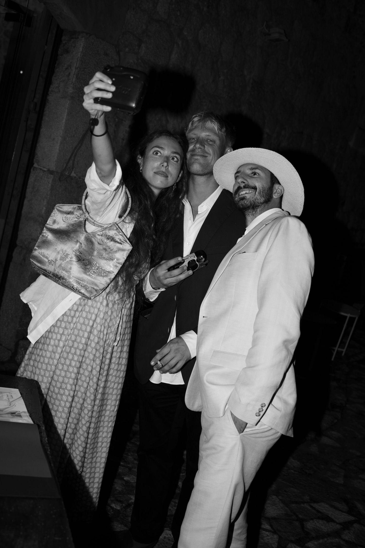 Black and white photo of three friends in stylish outfits posing for a night selfie, one wearing a white suit and wide-brim hat.