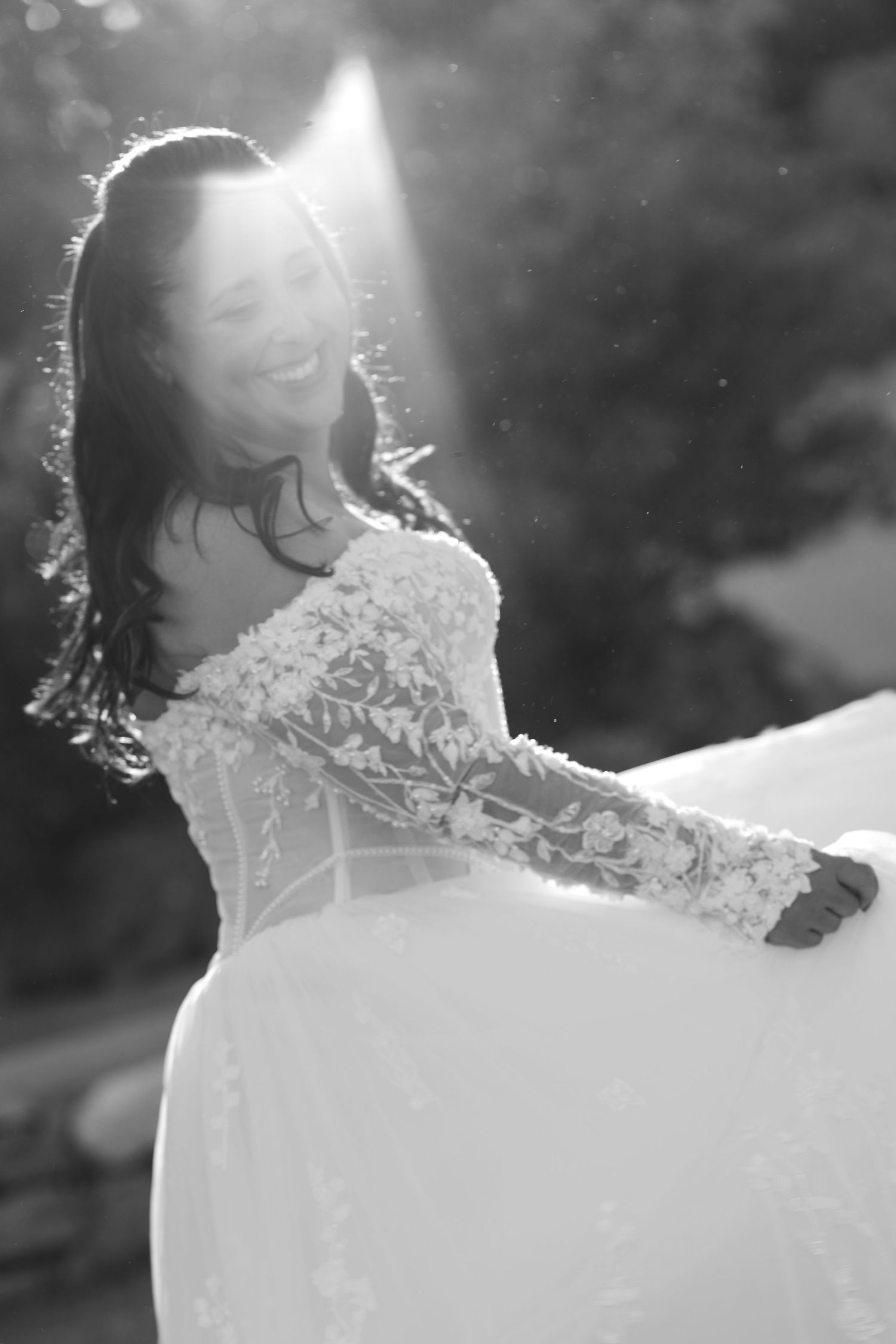 Smiling bride in a lace long-sleeve wedding gown twirls in glowing backlight in a black and white outdoor wedding portrait.